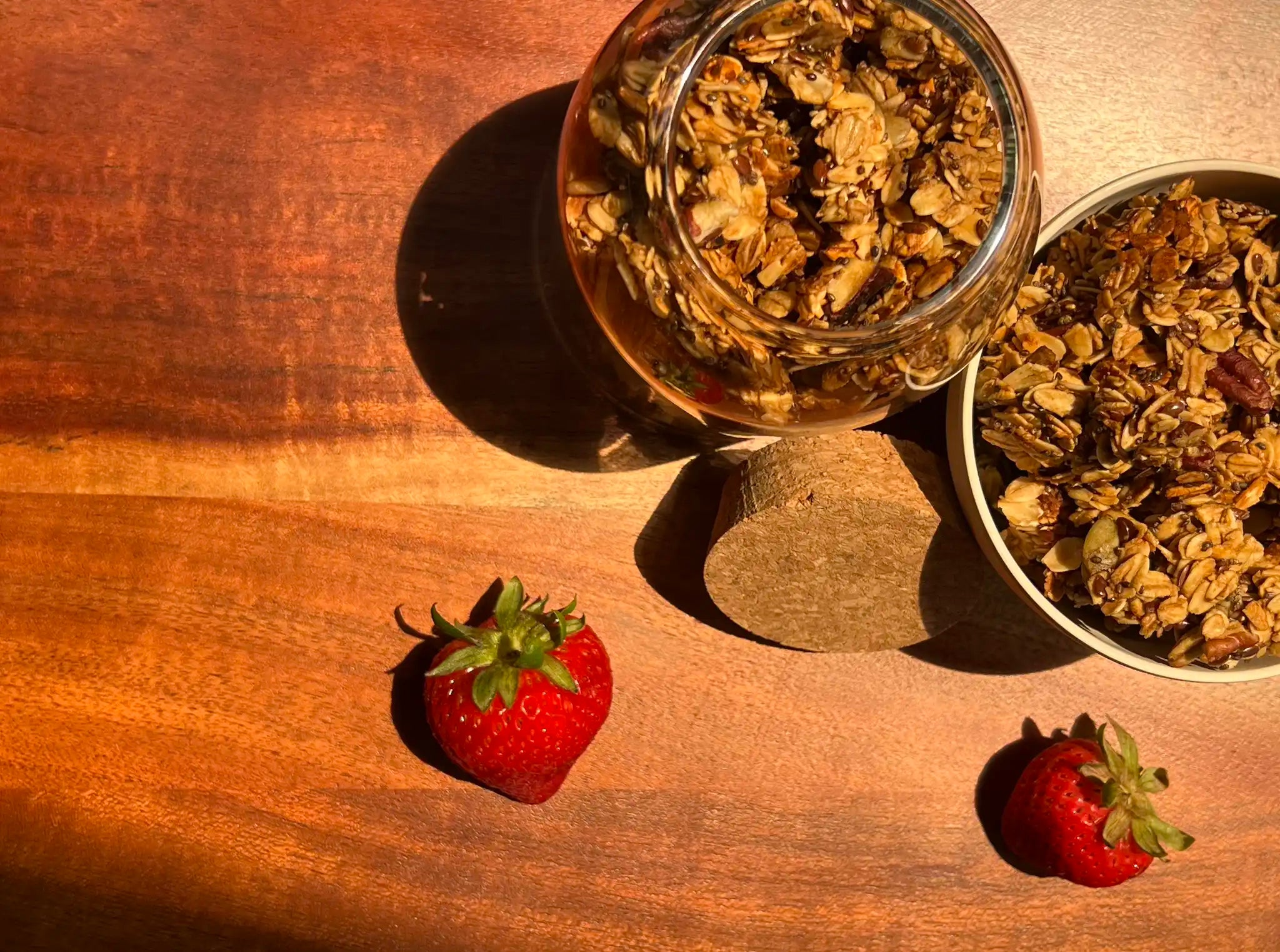 granola chunks in a glass jar and white bowl on a wooden background with scattered strawberries