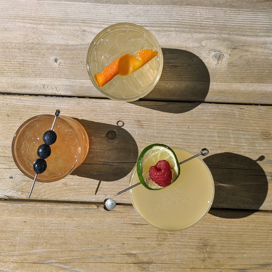 top down view of three cocktails garnished from left to right with blueberries, an orange twist, and a skewer with lime and a raspberry