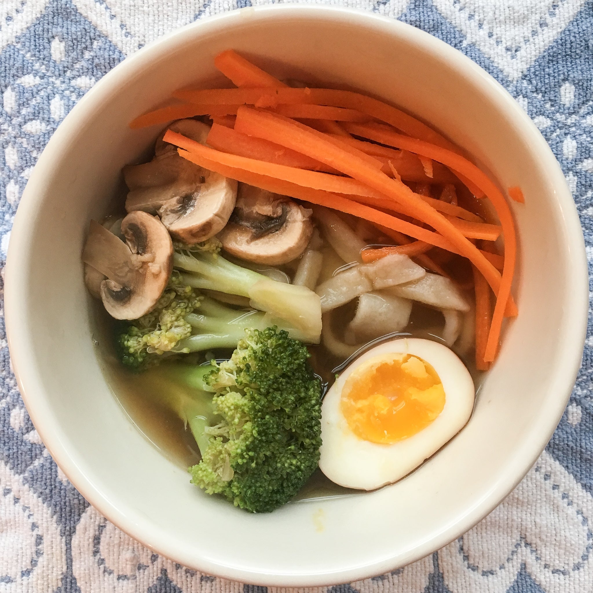 bowl with broccoli, mushrooms, sliced carrots, a hardboiled egg, and wood dragon udon broth on a blue and white tablecloth