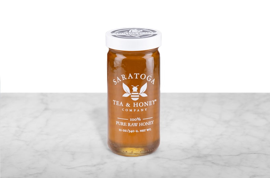 Amber-hued hot honey infused with Caroline Reaper pepper in a 12oz clear glass jar branded with the Saratoga Tea & Honey Co. logo and a white top. The logo and the words: "100% Pure Raw Honey" are printed on the jar.