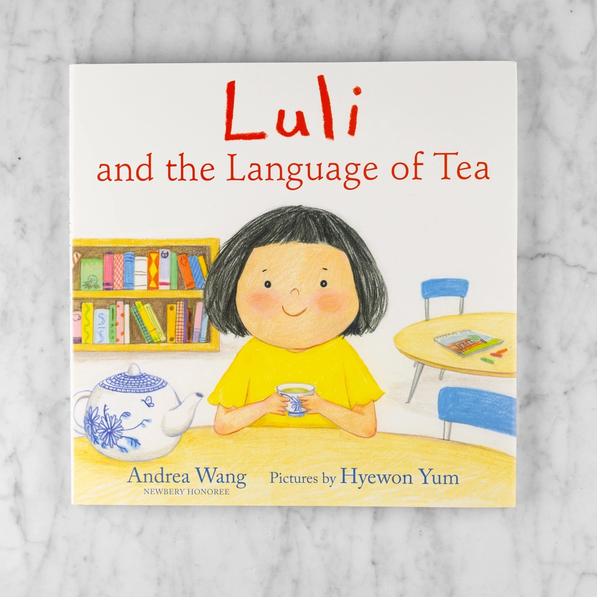Book cover of 'Luli and the Language of Tea' on a marble surface. The cover shows a little girl with black hair in a classroom drinking tea. Title: Luli and the Language of Tea. Author: Andrea Wang (Newberry Nominee). Pictures by: Hyewon Yum.