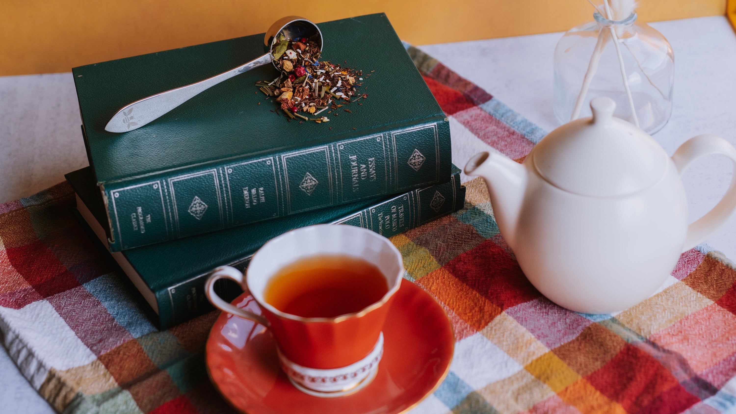 white teapot and orange vintage teacup with tea in front of a stack of old fashioned books with a tea scoop spilling tea on top