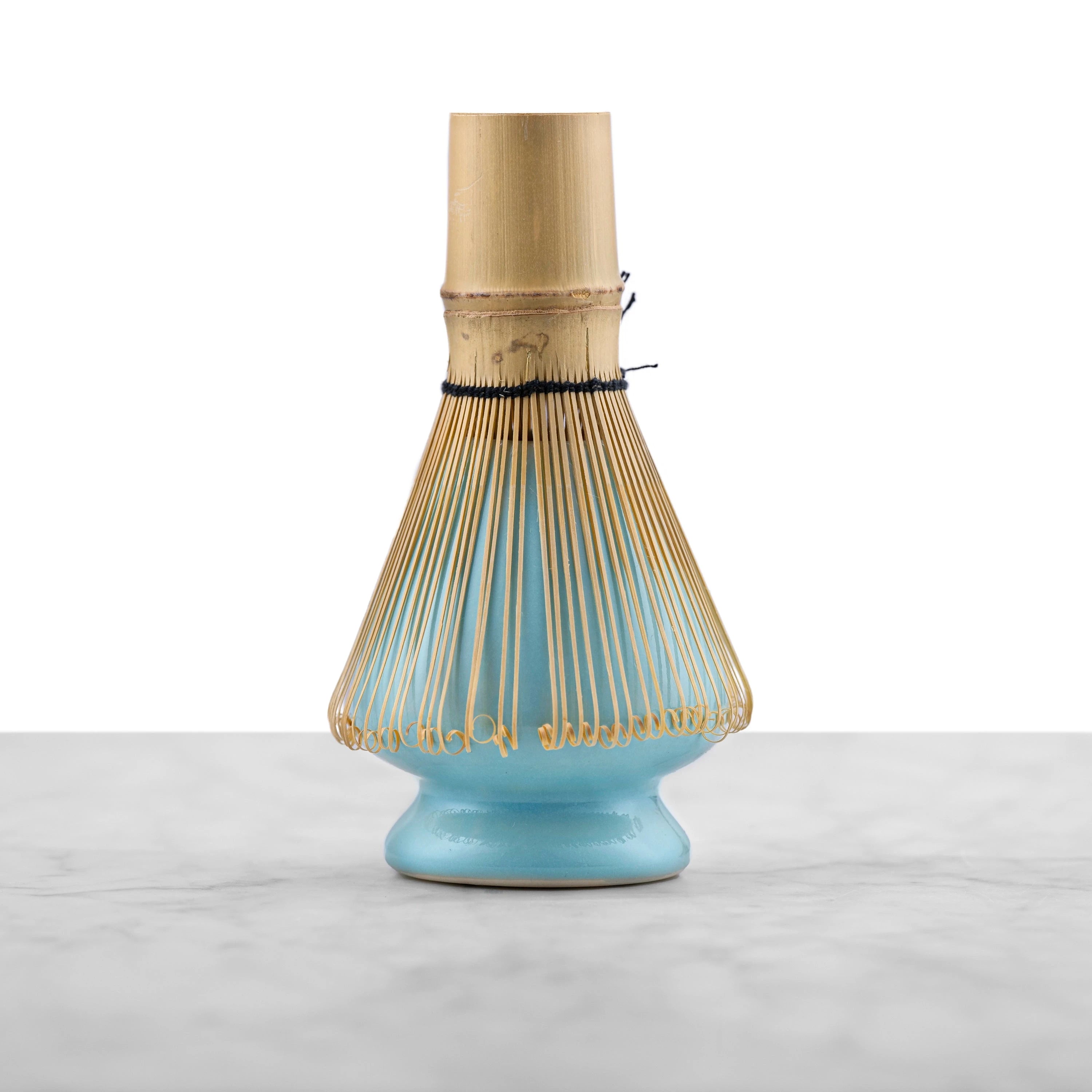 bamboo matcha whisk placed on top of a aqua colored ceramic matcha whisk stand in an tapered, rounded pyramid shape with a hole in the top center for the inner tines of the matcha whisk