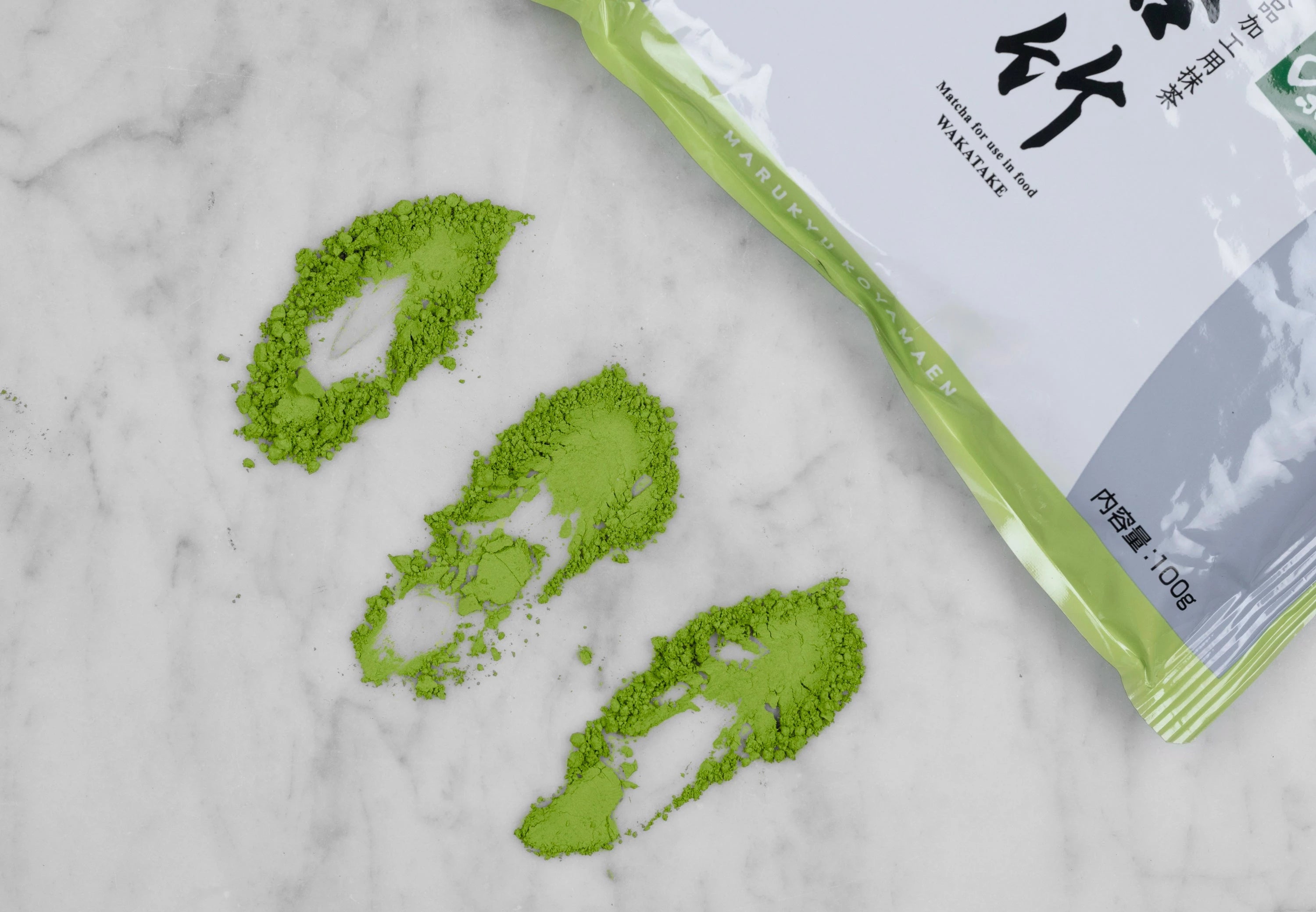Matcha Wakatake green tea power in three piles that have been drug towards the bottom left corner to show quality. A 100g pouch is displayed kitty-corner in the top right corner.