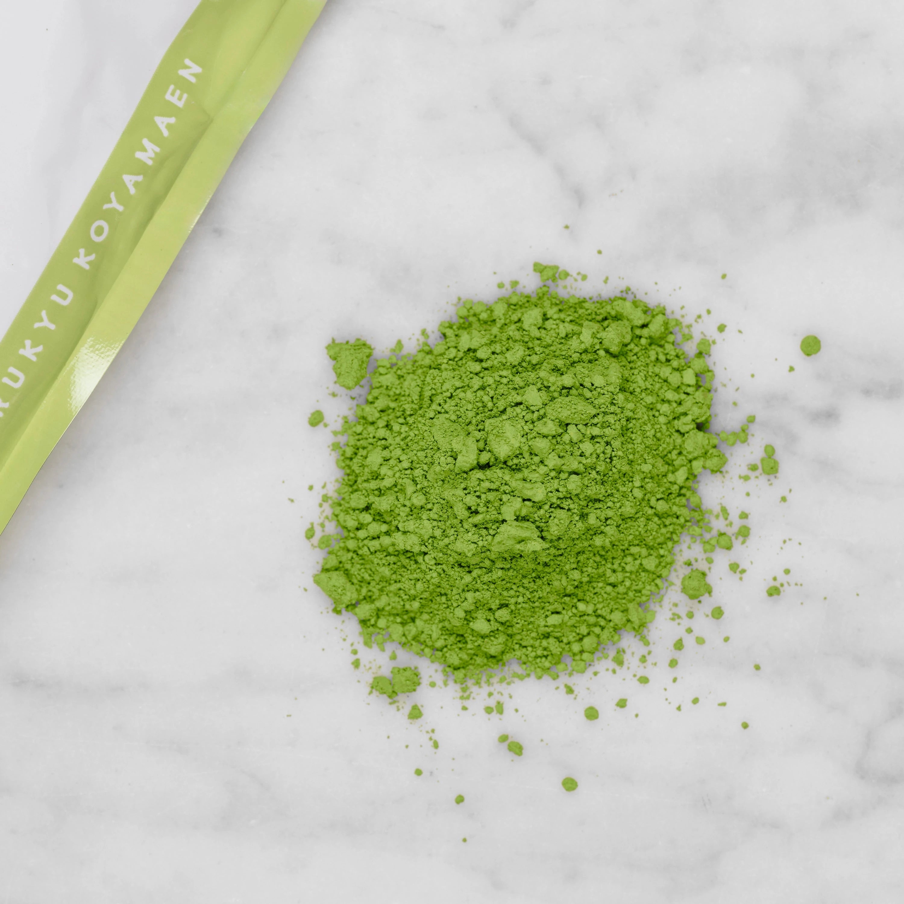 Matcha Wakatake green tea powder in a centered pile with a small corner of the 100g pouch reading "Marukyu Koyamaen" in the upper left corner.