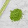 Matcha Wakatake green tea powder in a centered pile with a small corner of the 100g pouch reading "Marukyu Koyamaen" in the upper left corner.