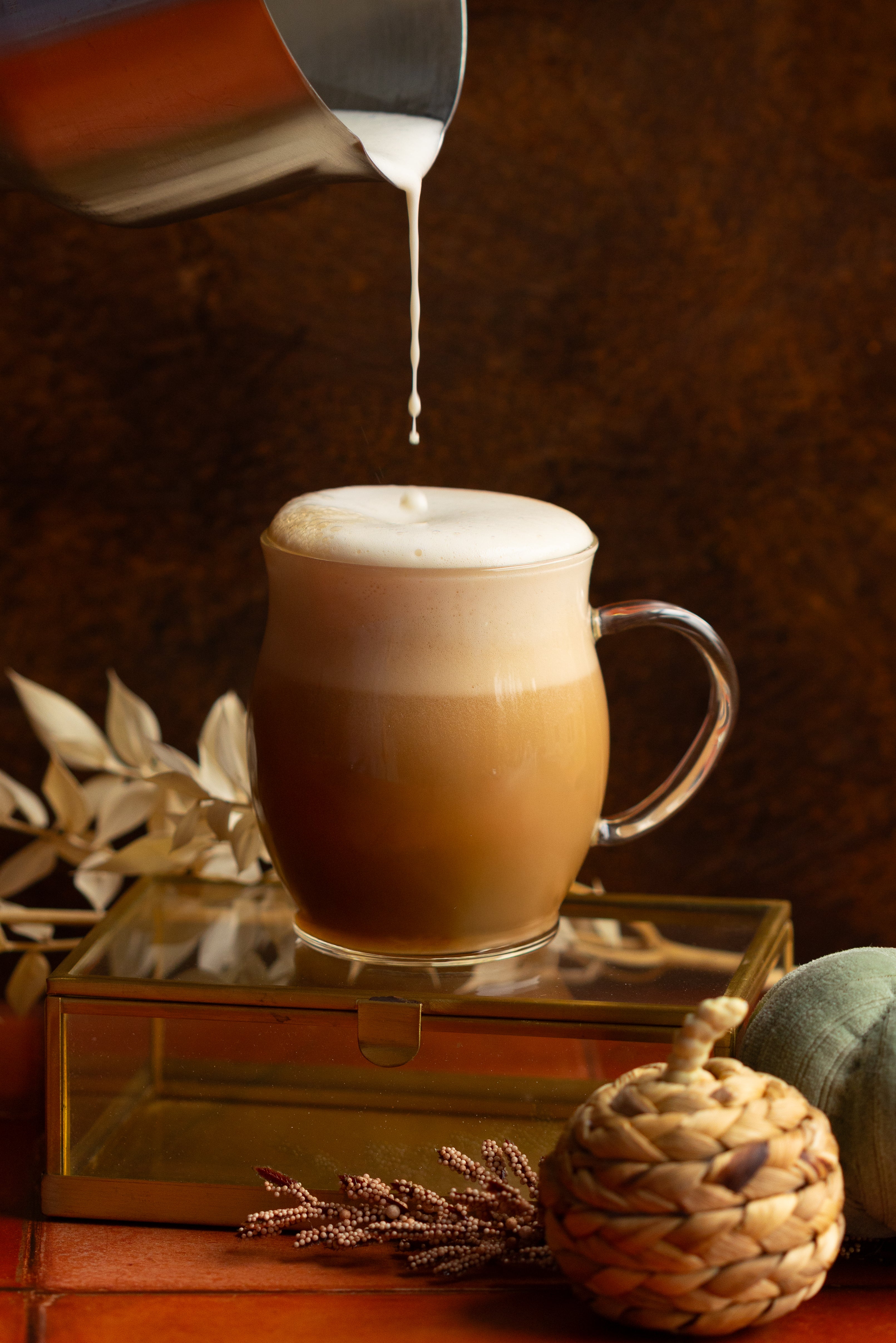 Cozy aesthetic scene of milk dripping into a very full chai latte in a glass mug. Scene is set with braided grass pumpkins, dried flowers, and moody lighting.