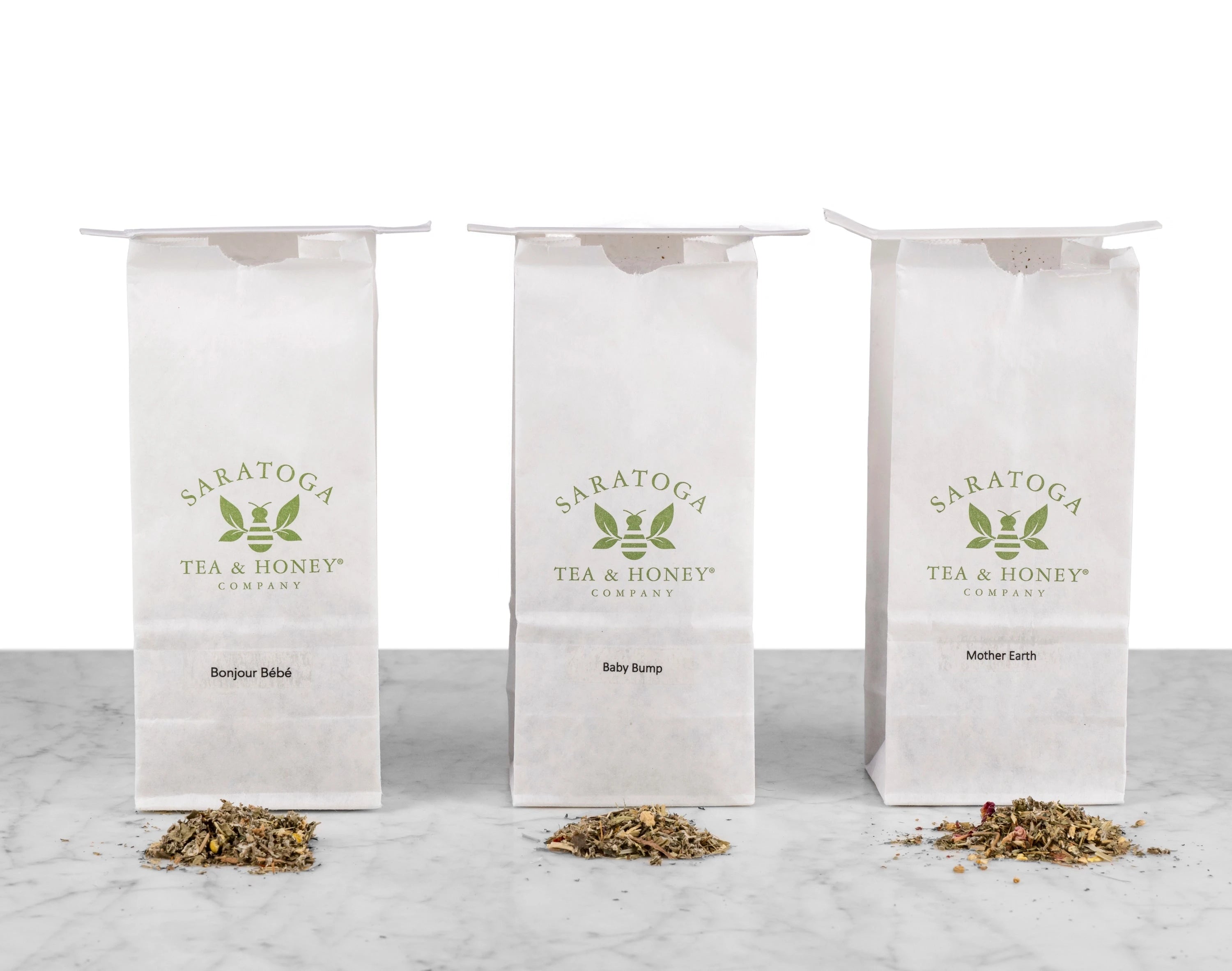 The three Motherhood & Fertility Bundle Teas lined up next to each other in white Saratoga Tea & Honey Co. tea bags with piles of corresponding teas in front: Bonjour Bebe, Baby Bump & Mother Earth