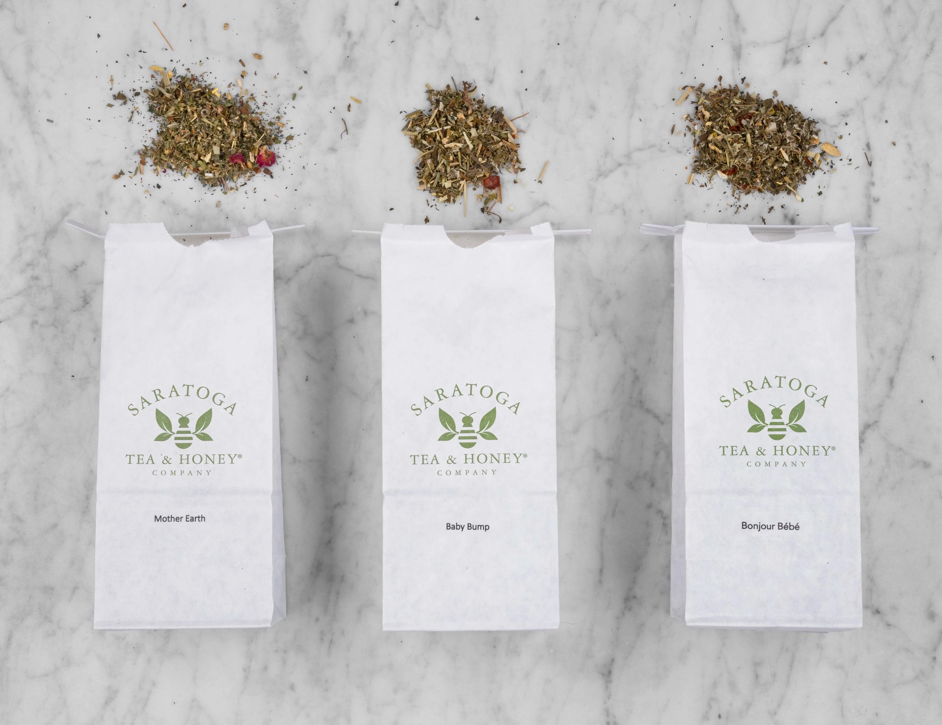 A flat lay of the three Motherhood & Fertility BundleTeas lined up next to each other  in white Saratoga Tea & Honey Co. tea bags with piles of corresponding teas at the top of the bags: Bonjour Bebe, Baby Bump & Mother Earth