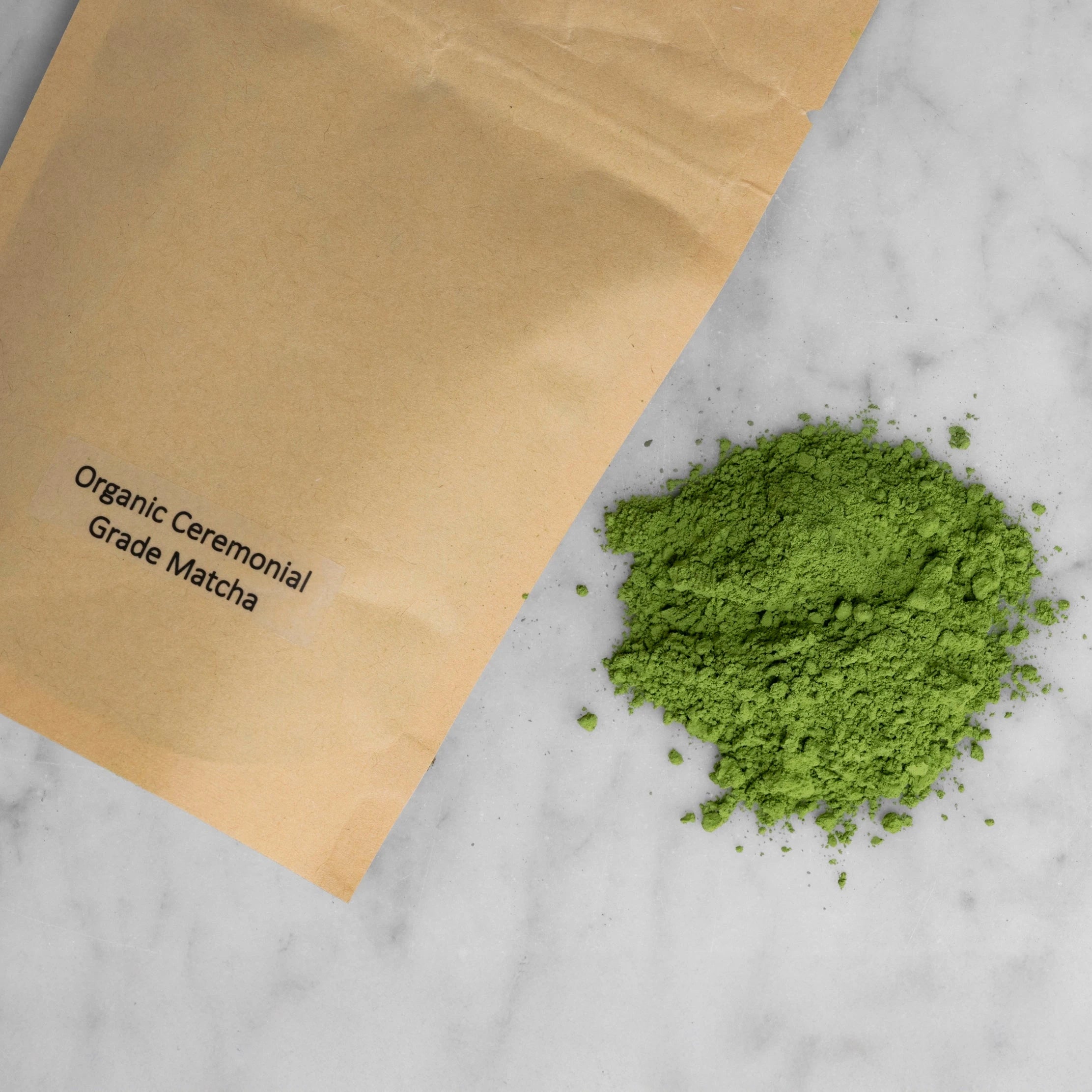 Packaged and unpackaged organic ceremonial grade matcha. A brown kraft paper resealable pouch lays next to a pile of vibrantly green organic ceremonial grade matcha from Japan