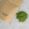 Packaged and unpackaged organic ceremonial grade matcha. A brown kraft paper resealable pouch lays next to a pile of vibrantly green organic ceremonial grade matcha from Japan
