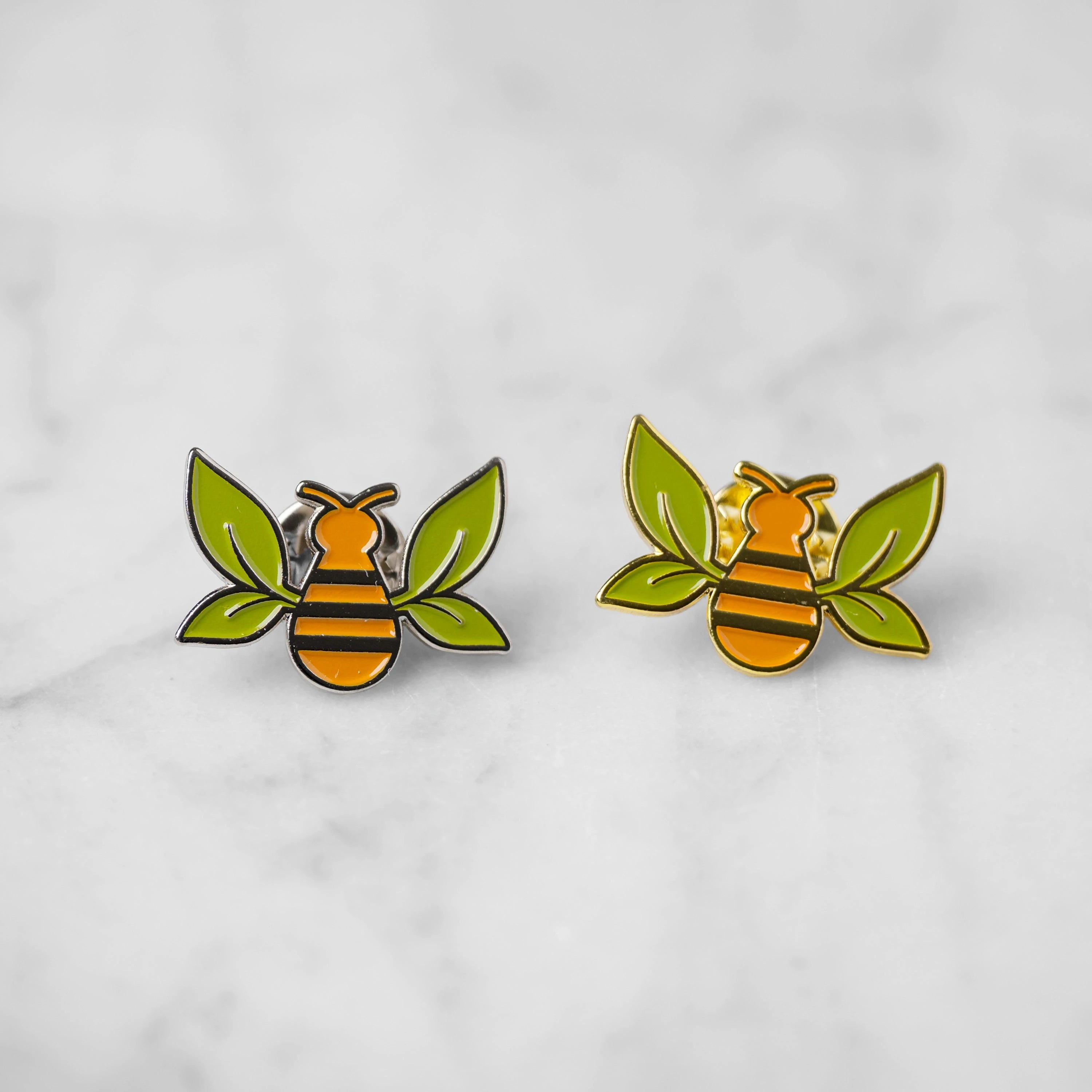 "Tea Bee" enamel pins with green tea leaf wings and orange bee bodies. Silver enamel on left and gold enamel on right.
