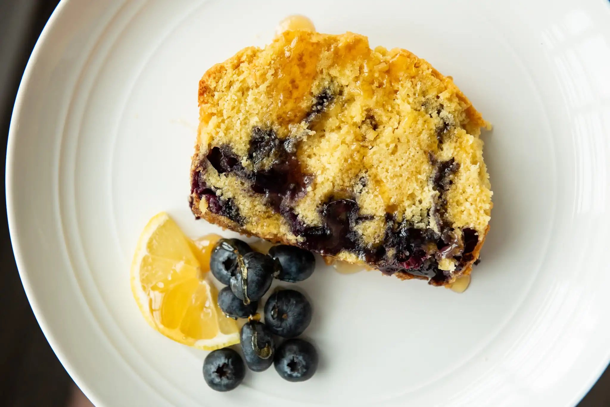 slice of blueberry ginger lemon tea bread on a white plate with blueberries and a lemon slice, all drizzled with honey
