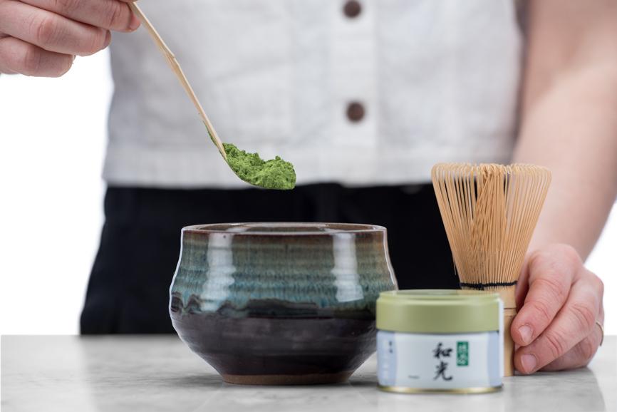 hand measuring bright green matcha into a matcha bowl and holding a matcha whisk