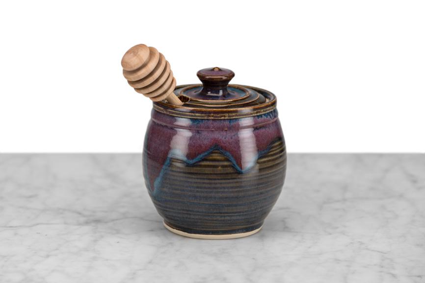 dark brown and purple clay honeypot with wooden honey dipper on a marble and white background