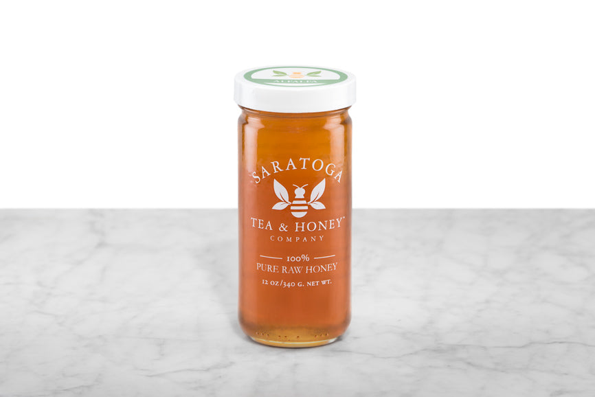 Amber-hued Alfalfa Honey displayed in a 12oz cylindrical glass jar with a white lid. The logo and the words: "100% Pure Raw Honey" are printed on the jar.