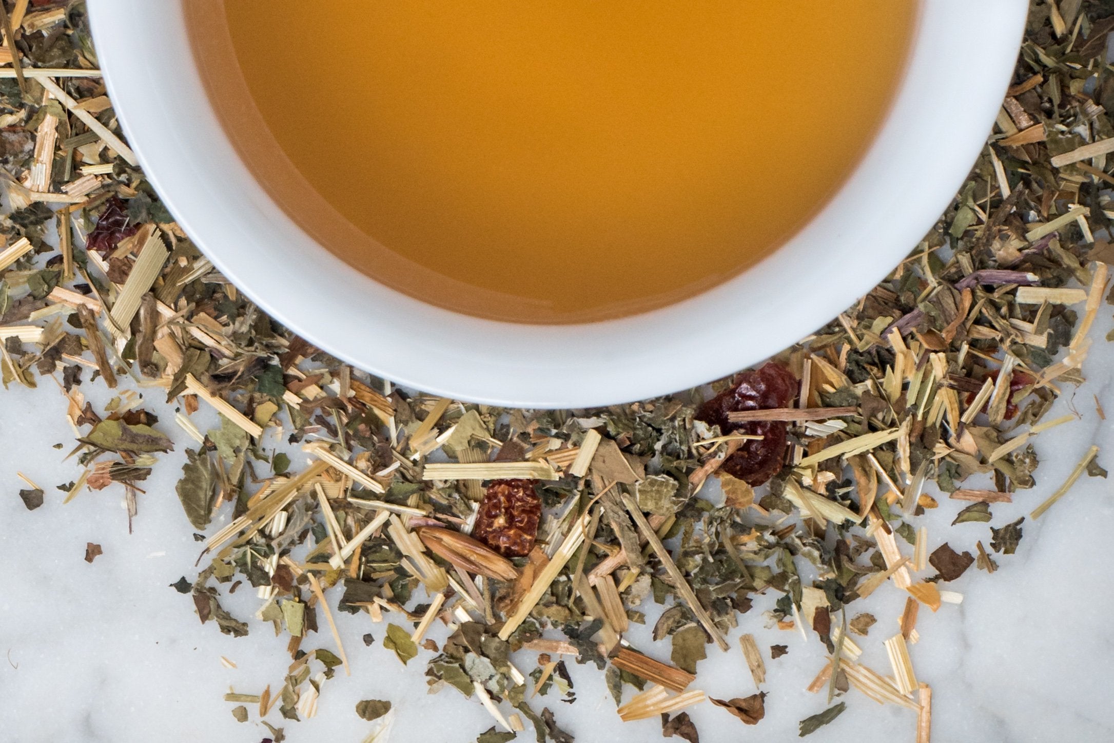 Baby Bump leaves and brewed tea. Torn oat straw, small green leaves and dried rose hips surround a cup of ochre-colored brewed tea.