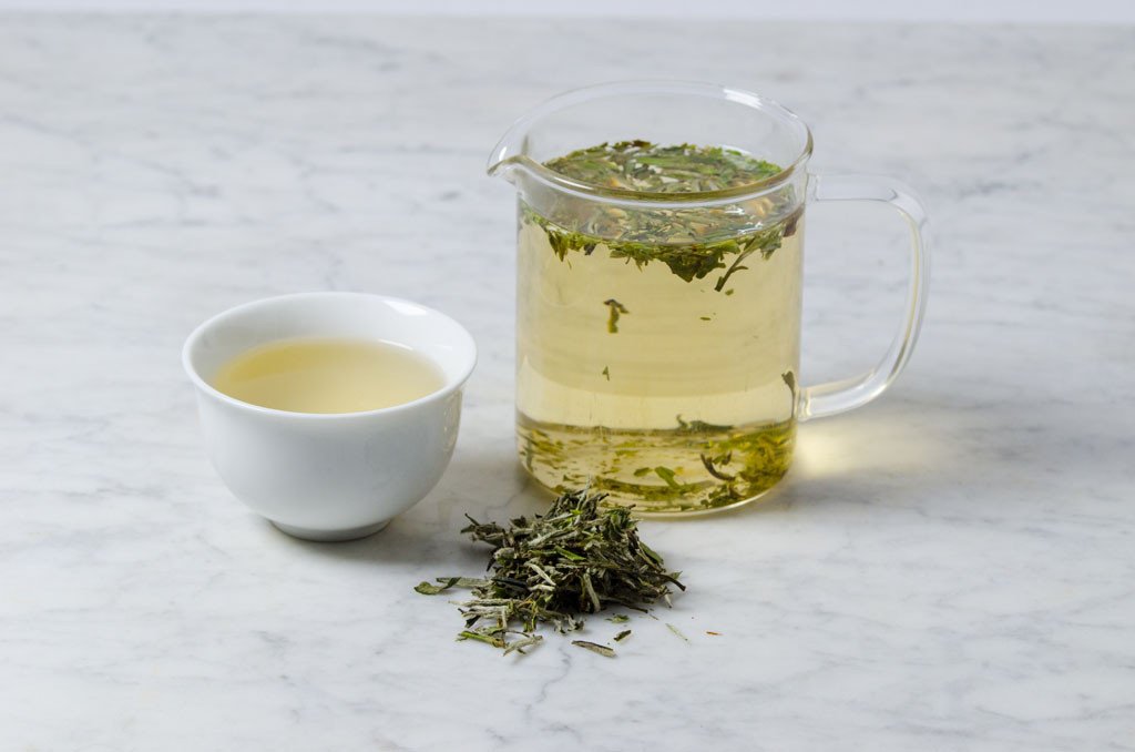 loose leaf bai mu dan wang brewing in a glass pitcher, in a white cup, and in a pile of dry leaves