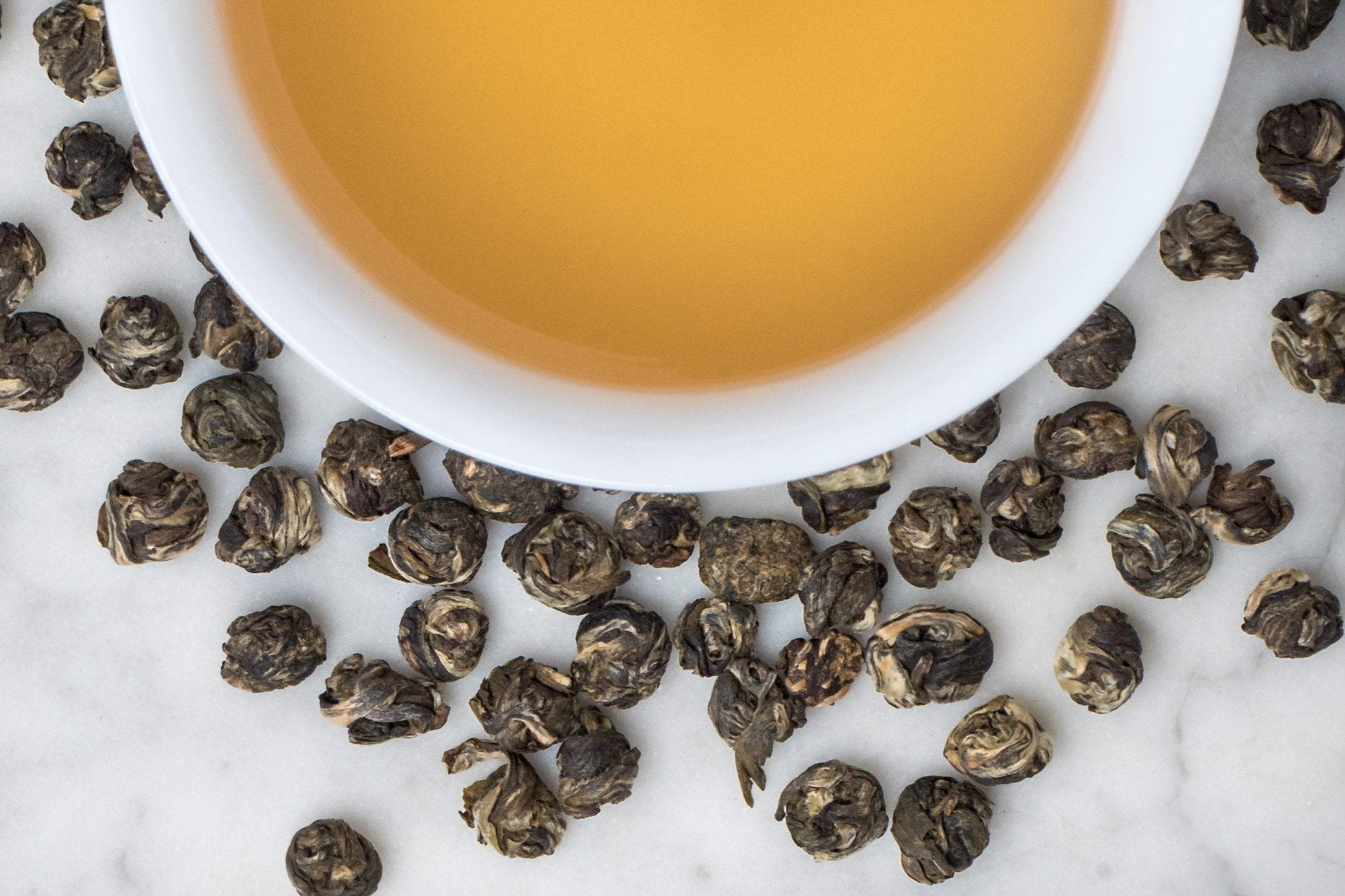 Jasmine Pearls leaves and brewed tea. Tightly rolled, small round balls of variagated green tea leaves surround a cup of honey-yellow brewed green tea.