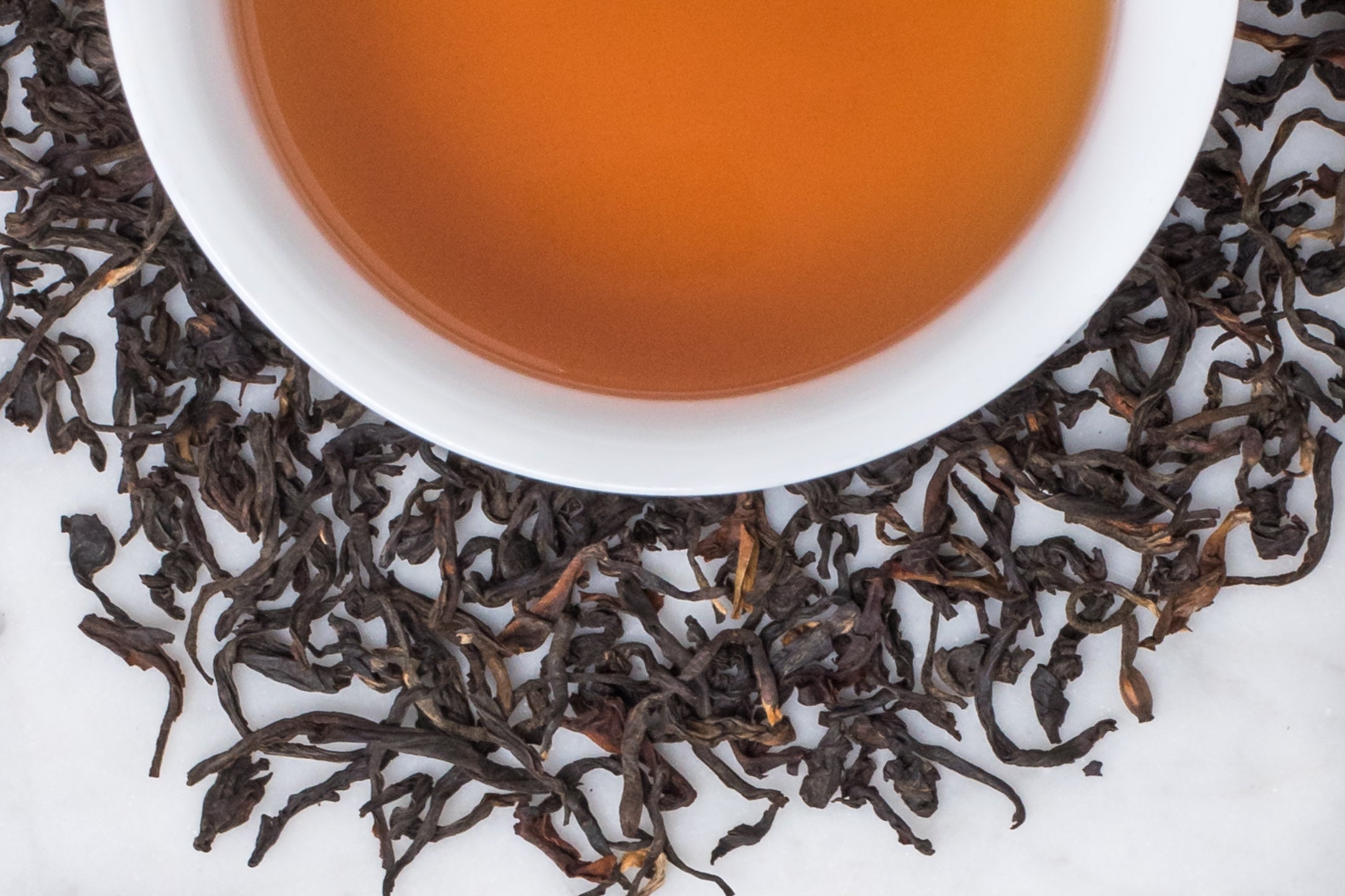 La Cumbre Bitaco leaves and brewed tea. Twisted dark brown and gold leaves surround an amber-hued cup of brewed black tea.