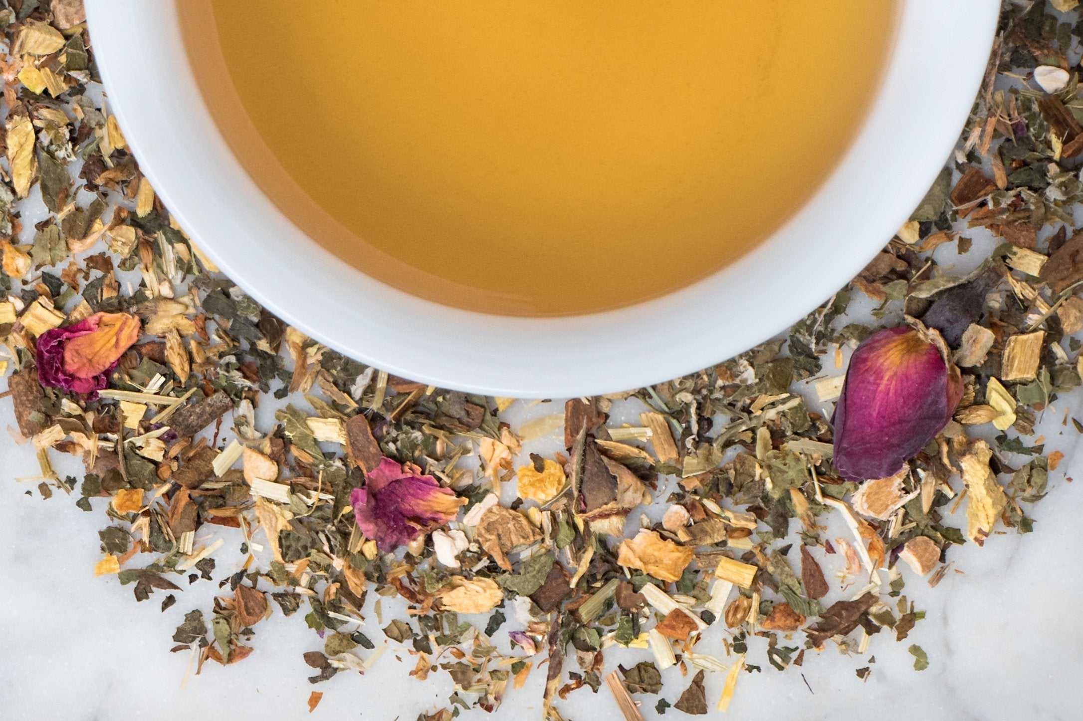 Mother Earth leaves and brewed tea. A mix of green, brown, and tan leaves with rose petals surrounds a cup of old-gold colored brewed tea.