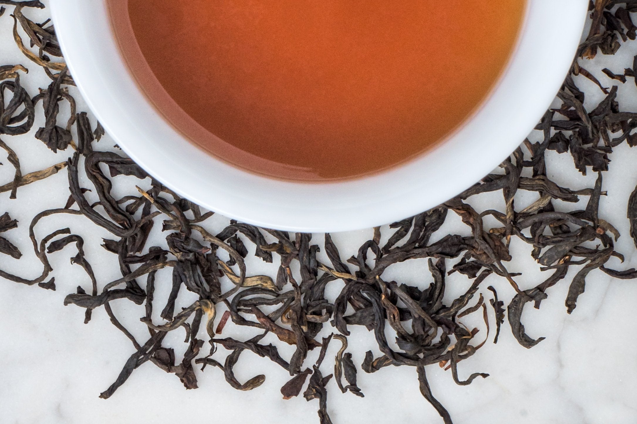 Russian Caravan leaves and brewed tea. Long and thin twisted black tea leaves surround a cup of dark-amber brewed black tea.