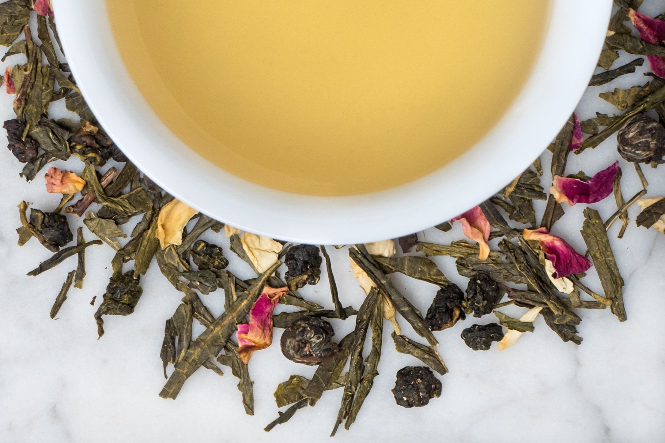Saratoga Wedding Tea leaves and brewed tea. A mix of long green tea leaves, rolled oolong leaves, and rose surround a cup of light yellow brewed tea.