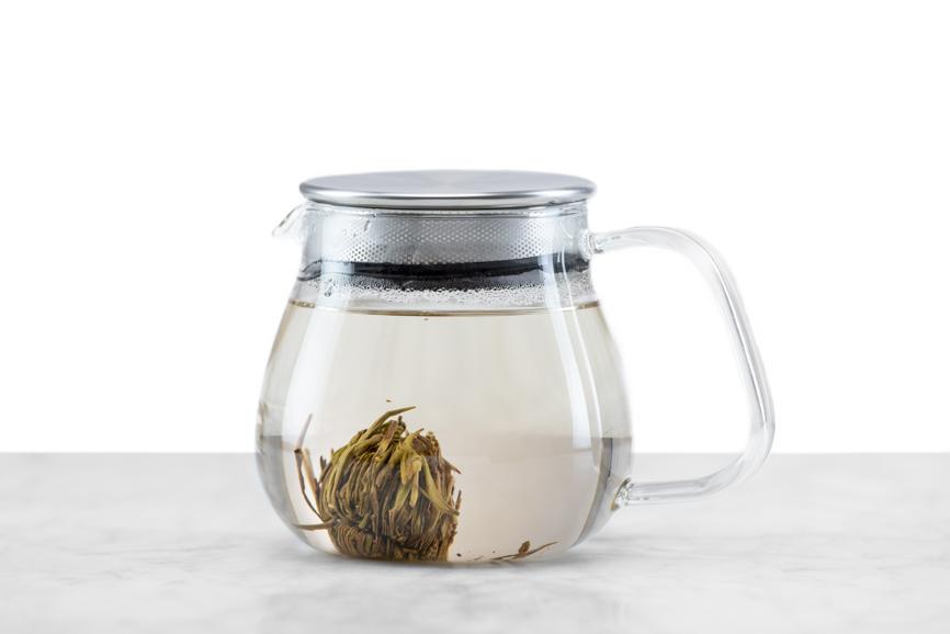 Jasmine Flower Craft tea ball brewing stage 3 - steeping part 2. Tea ball is resting on the bottom of the glass teapot and leaves are opening. The liquid in the teapot is starting to take on a yellow tinge from the tea