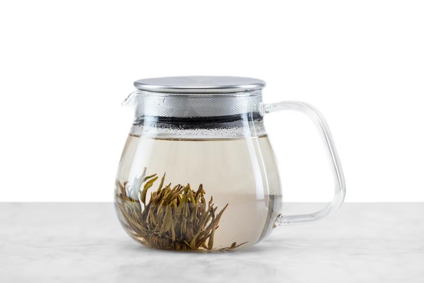 Jasmine Flower Craft tea ball brewing stage 4 - steeping part 3. Nearly open tea ball rests on the bottom of the glass teapot like a sea anemone with leaves reaching out into the water. The tea is definitely starting to take on a distinct hue.