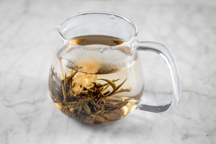 Jasmine Flower Craft blooming tea aerial view. Tea ball is fully open with unfurled jasmine blooms submerged in clear, light gold liquid.