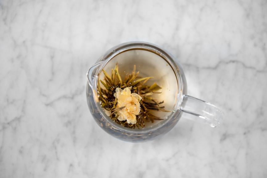 Jasmine Flower Craft blooming tea fully aerial view of open flower-like tea ball steeping in a glass teapot.