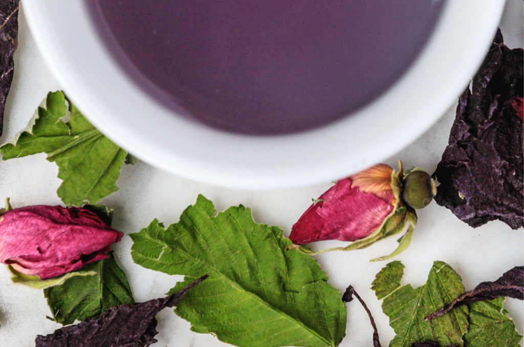 La Vie En Rose leaves and brewed tea. Purple leaves, green leaves, and full pink rosebuds surround a cup of dark purple brewed herbal tea.