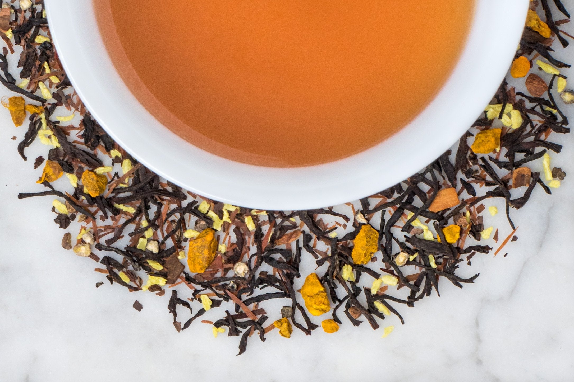 Vitality leaves and brewed tea. Twisted black tea leaves, coconut pieces, cinnamon and turmeric surround a cup of russet-hued brewed tea.