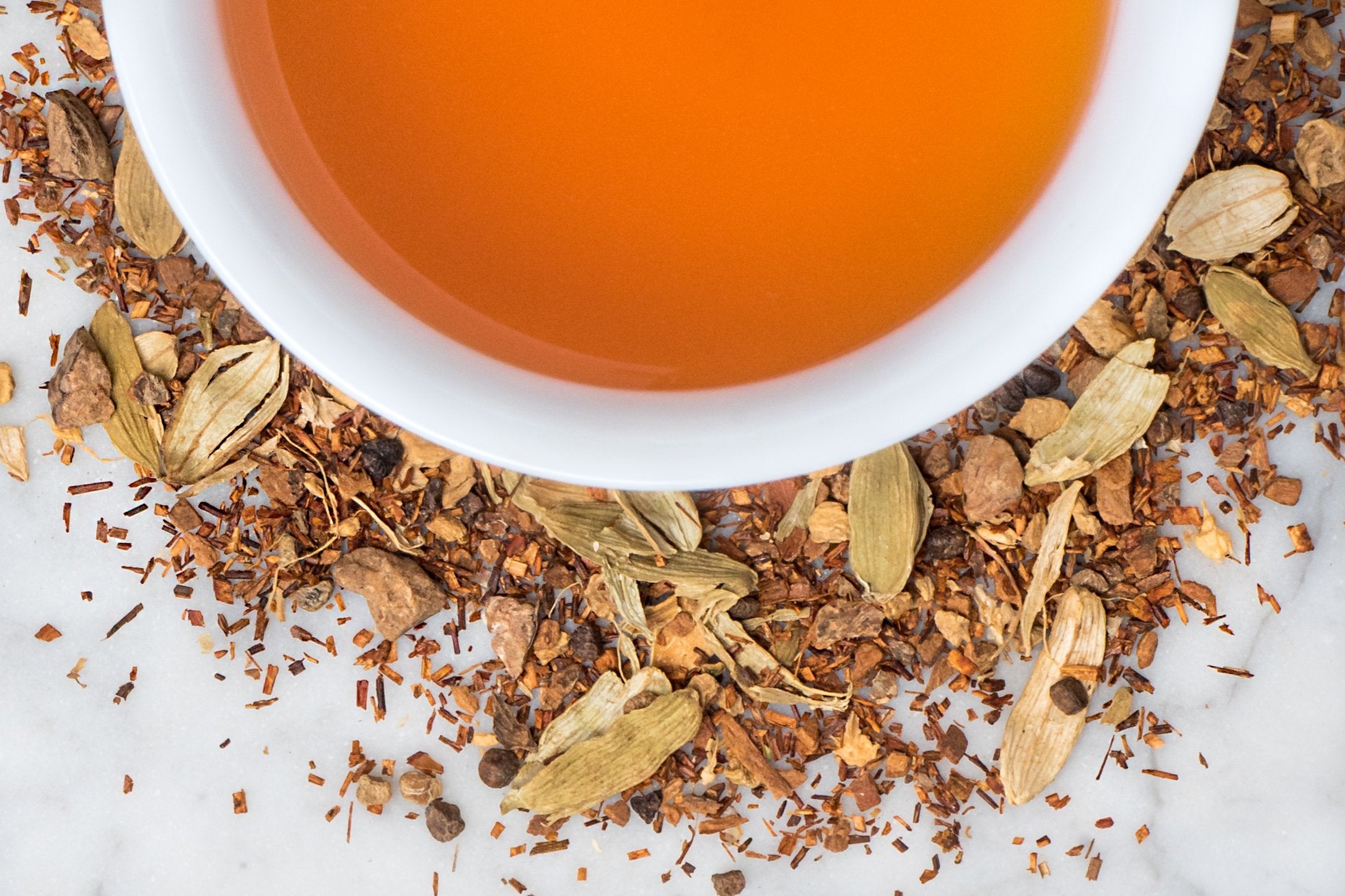 Saratoga Red Chai leaves and brewed tea. Fine pieces of rooibos, spices, and cardamom pods surround a cup of orangey-brown brewed tea.