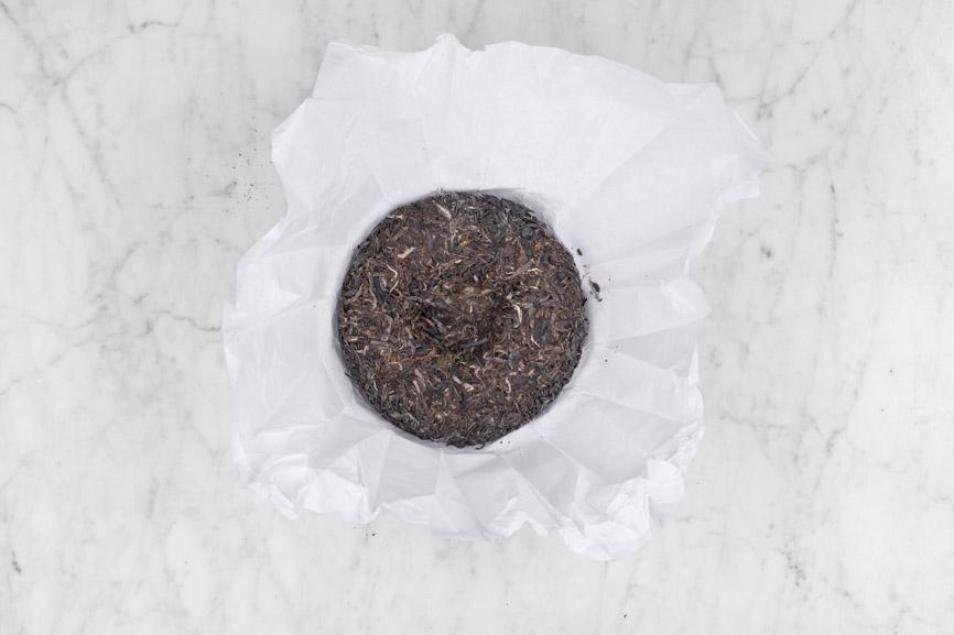 2007 Yiwu Jing Long sheng pu'er full unwrapped pu erh cake sitting in paper. The round cake is composed of brownish grey leaves and buds compressed in a random pattern of leaves.