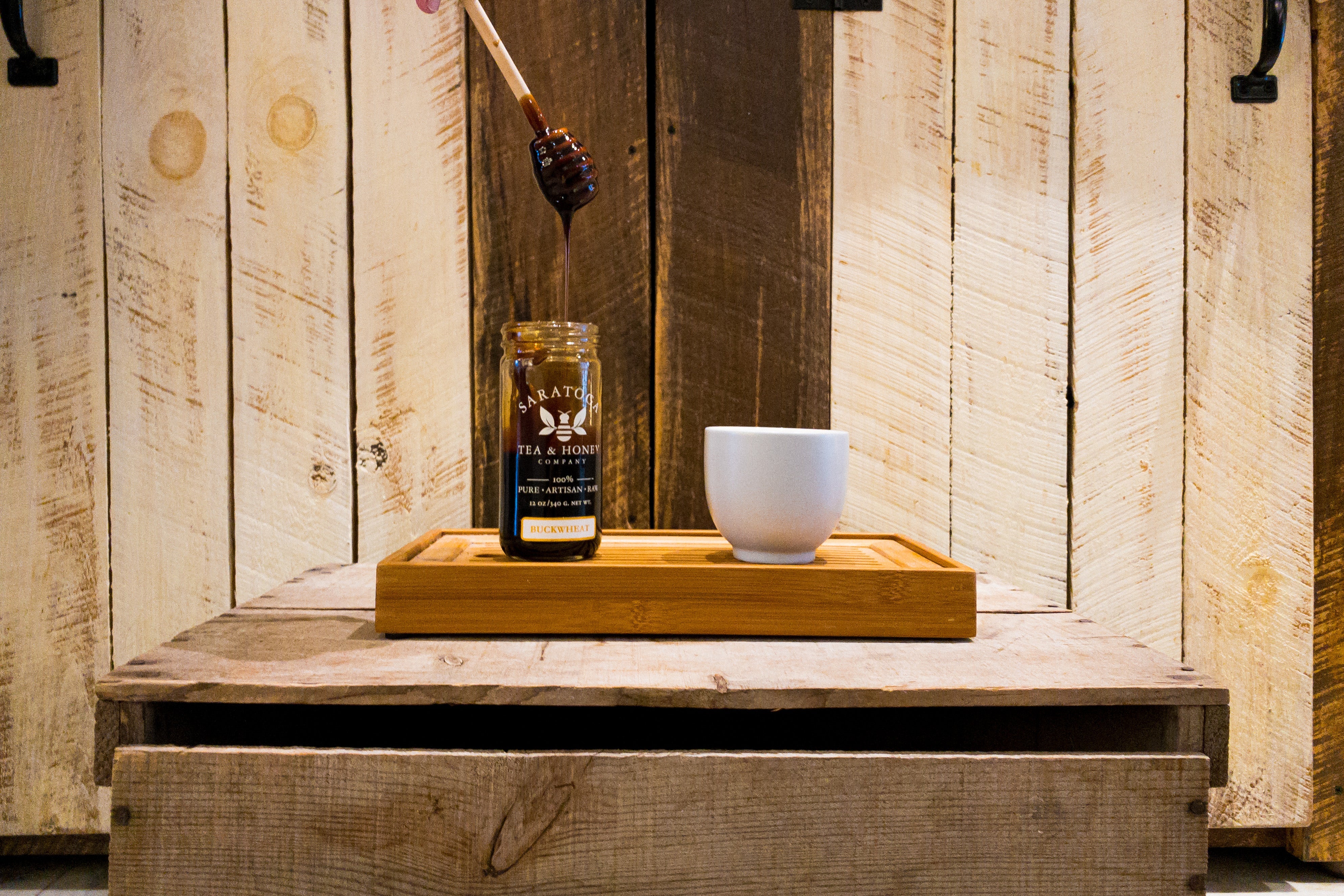 honey dipper dripping dark buckwheat honey into a jar of Saratoga Tea & Honey Co. dark honey sitting on a tea boat next to a handleless cup of tea
