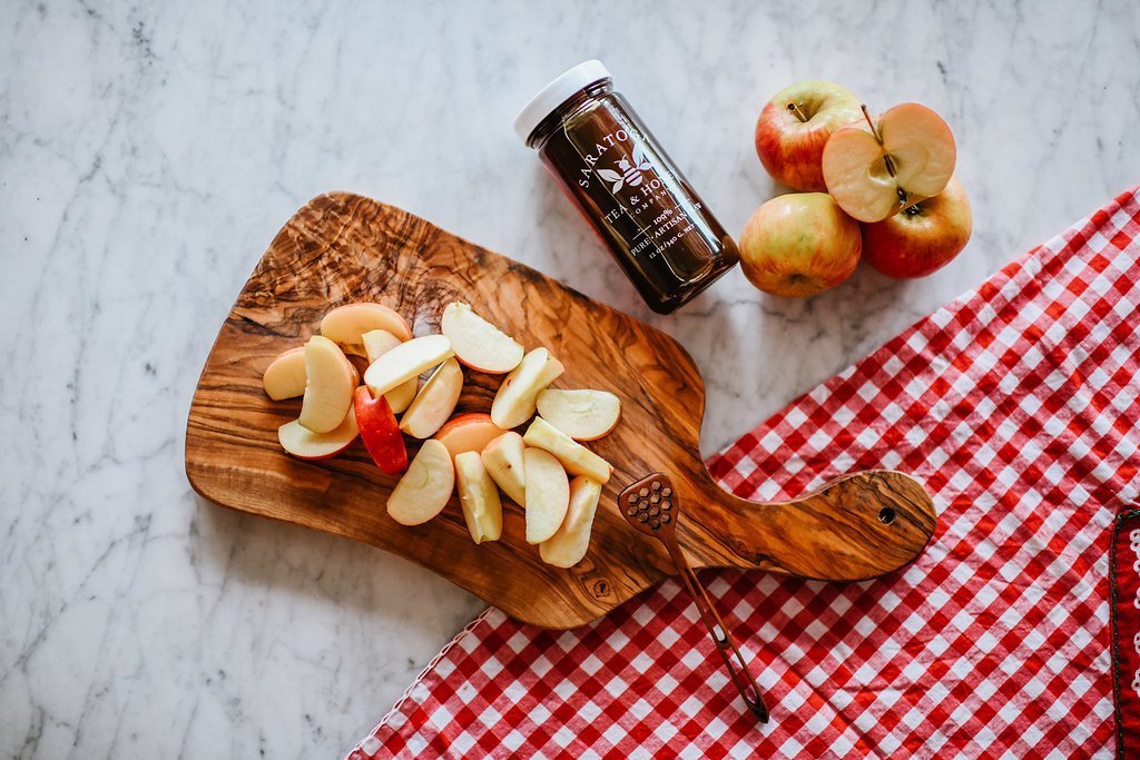 small wooden board with sliced apples next to a jar of cinnamon honey and a pile of apples 