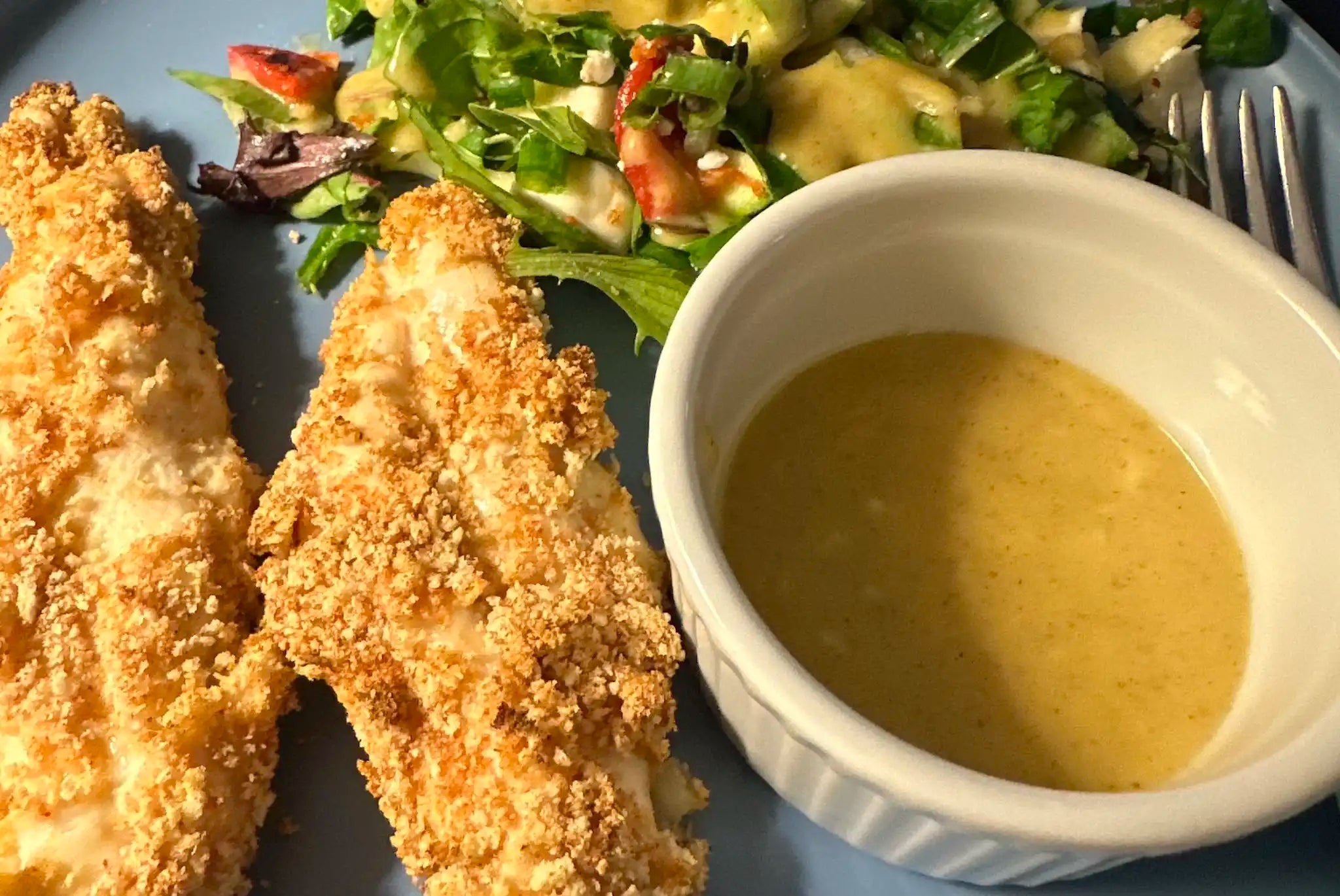fried chicken tenders next to a white ramekin of yellow hot mango honey mustard with a lightly dressed salad in the background