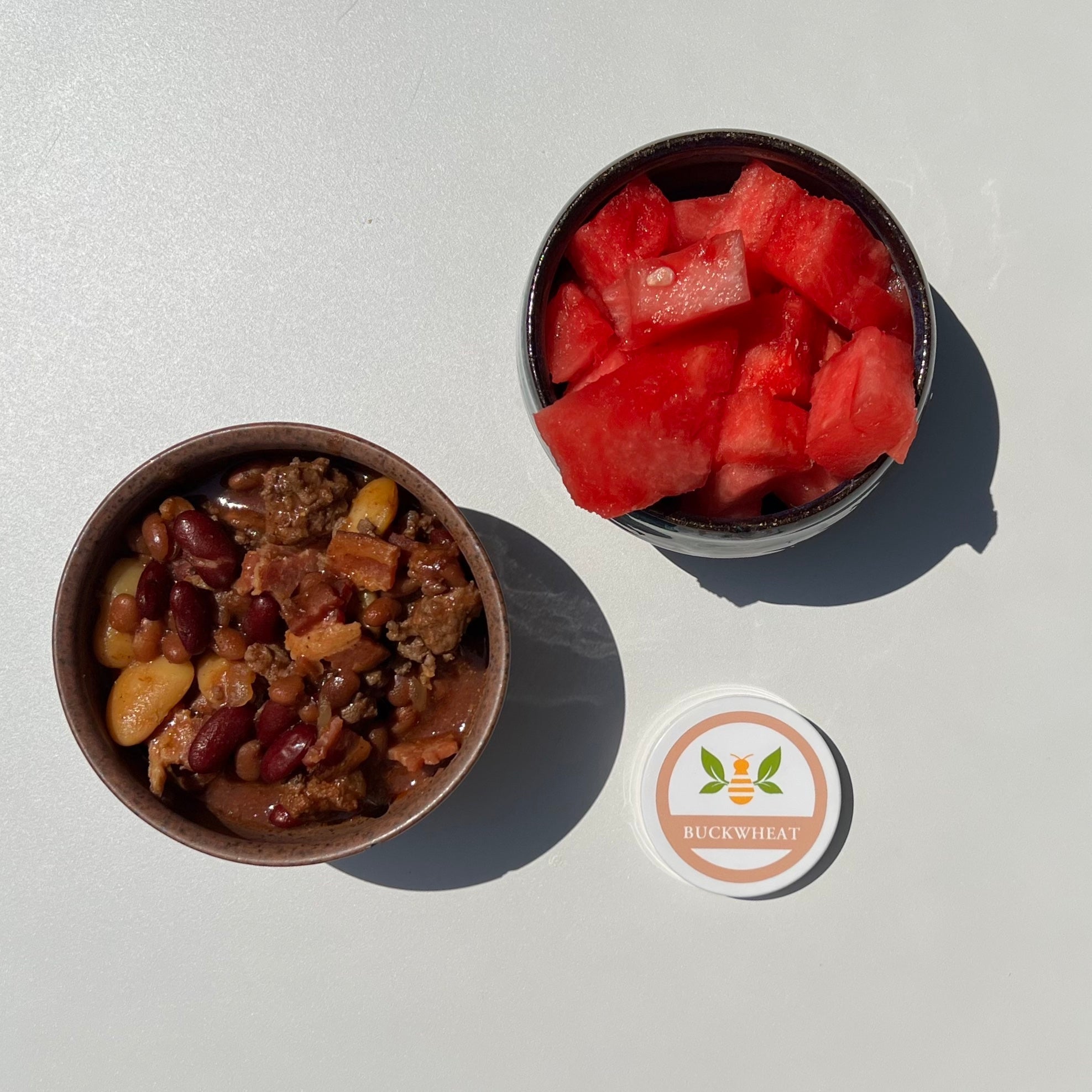 small brown bowl of barbeque baked beans next to a small bowl of watermelon and a lid for Saratoga Tea & Honey co. buckwheat honey