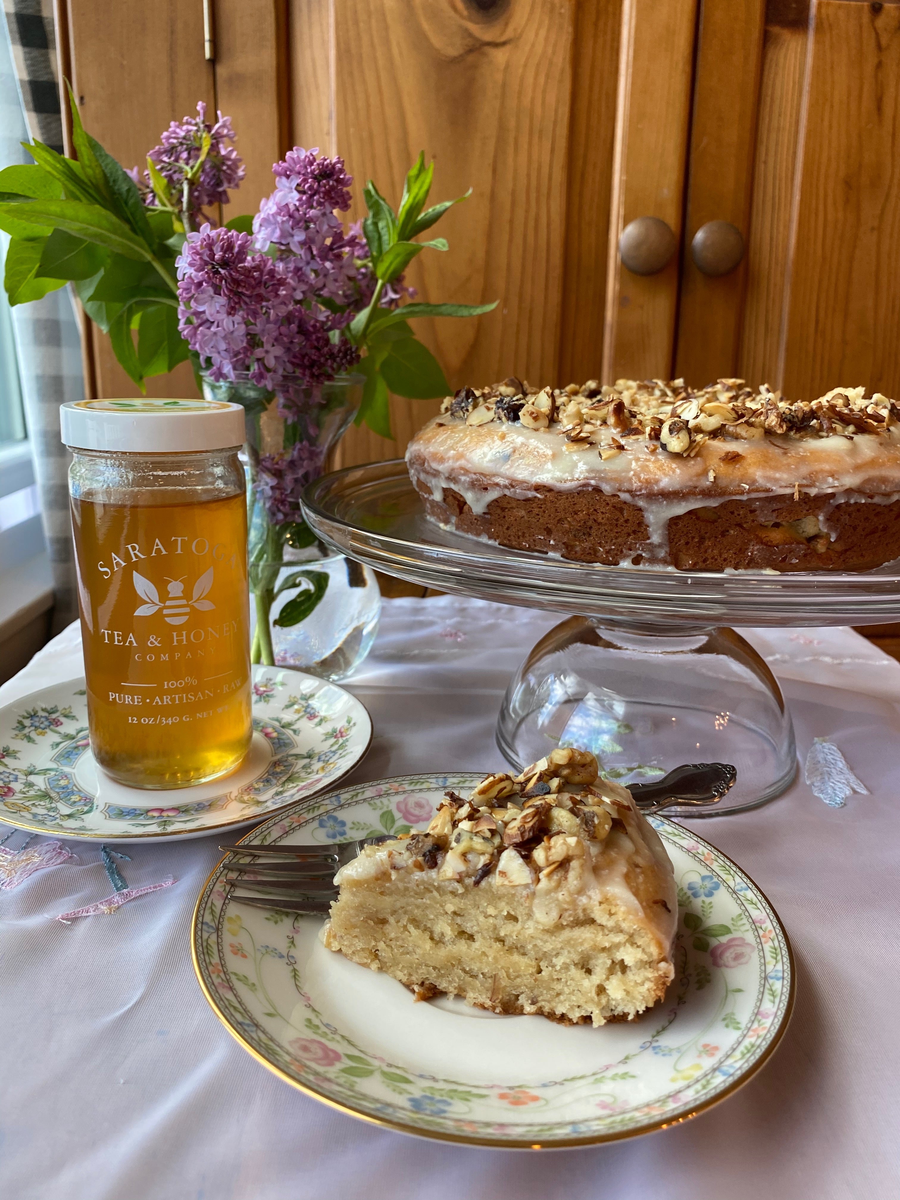 glazed coffee cake on a glass cake stand with a small vintage plate of cake and a jar of honey