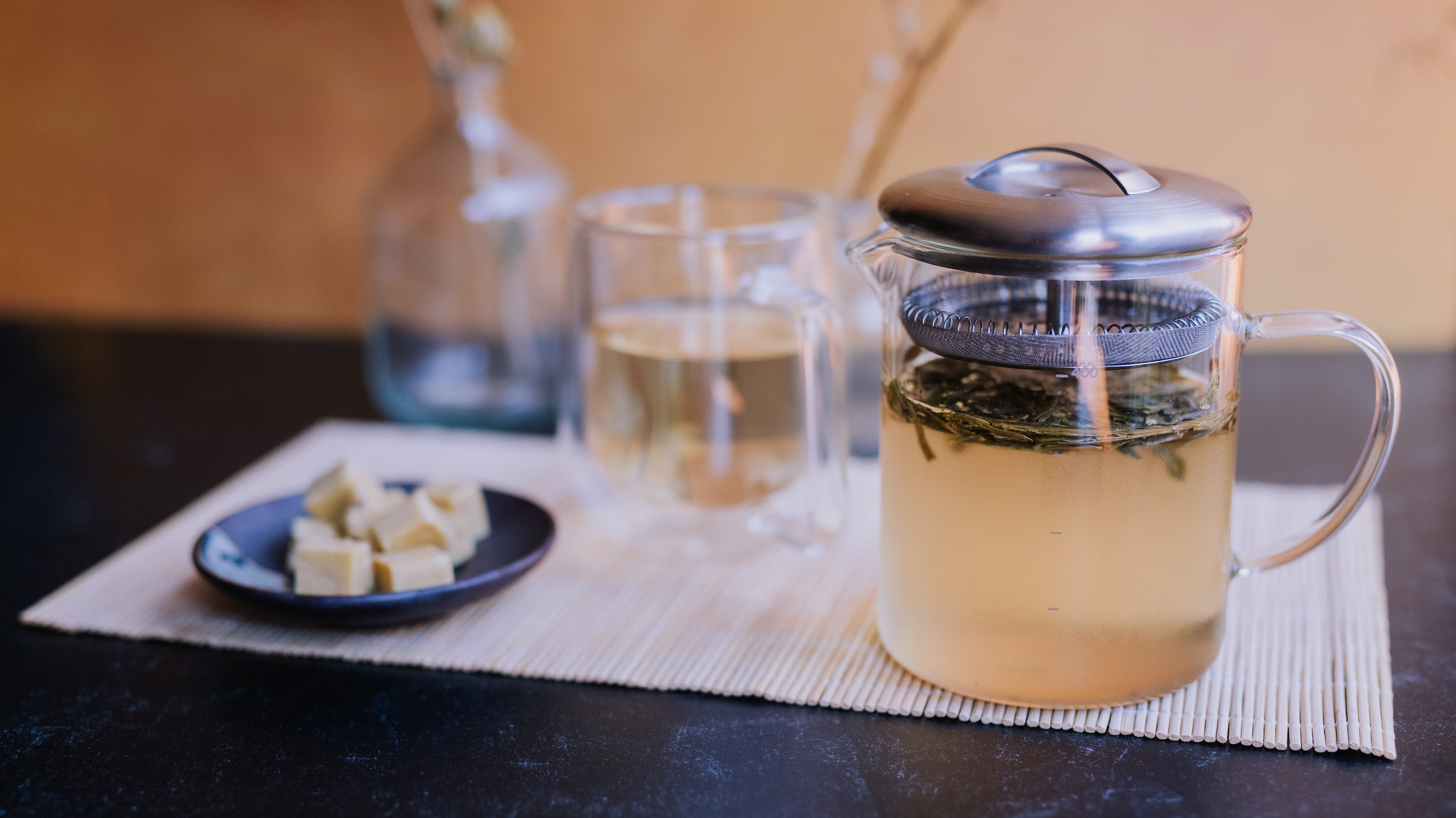 glass infuser of green tea next to glass mug of green tea and plate of mung bean cakes