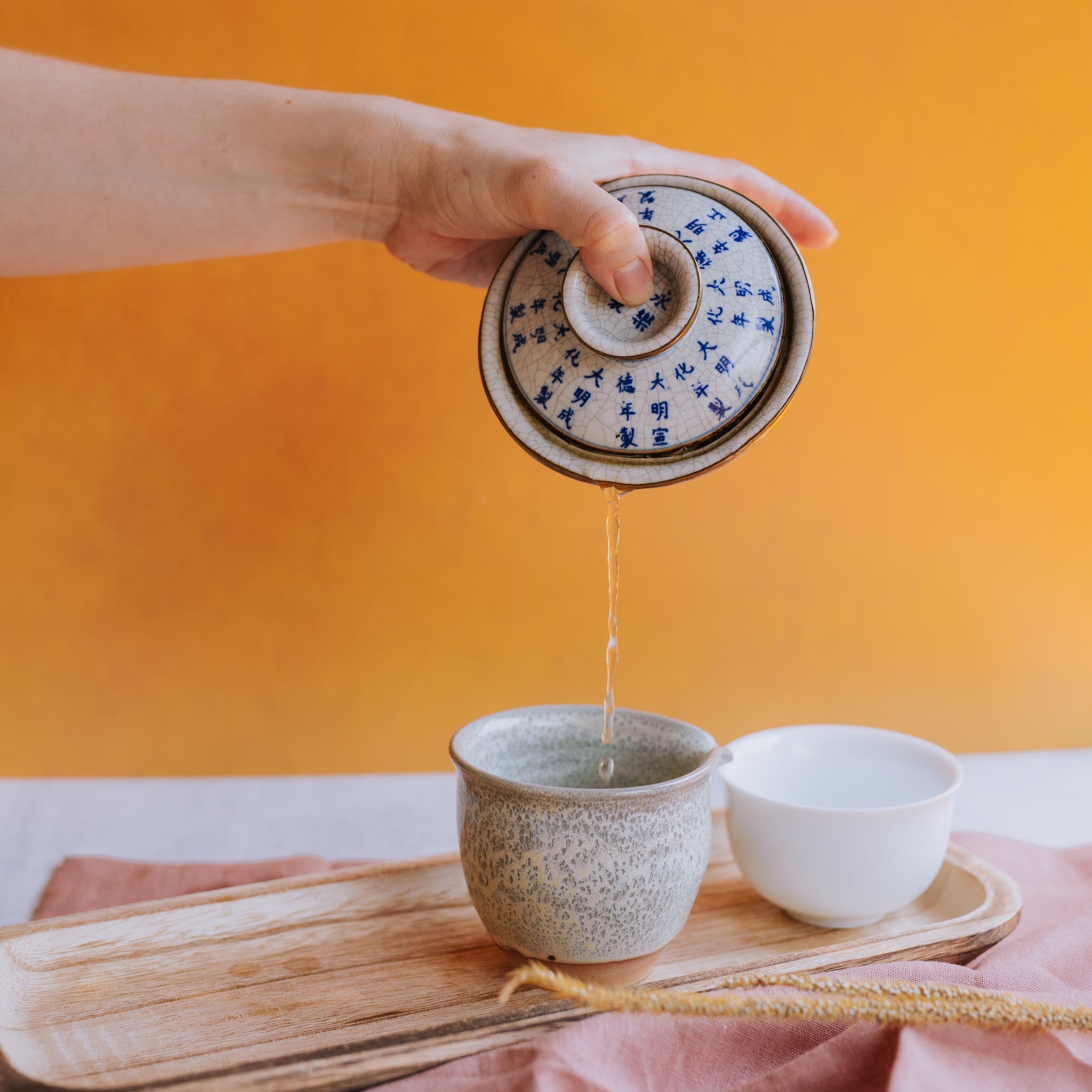 hand pouring white tea from a gaiwan into a pitcher against a mustard yellow background