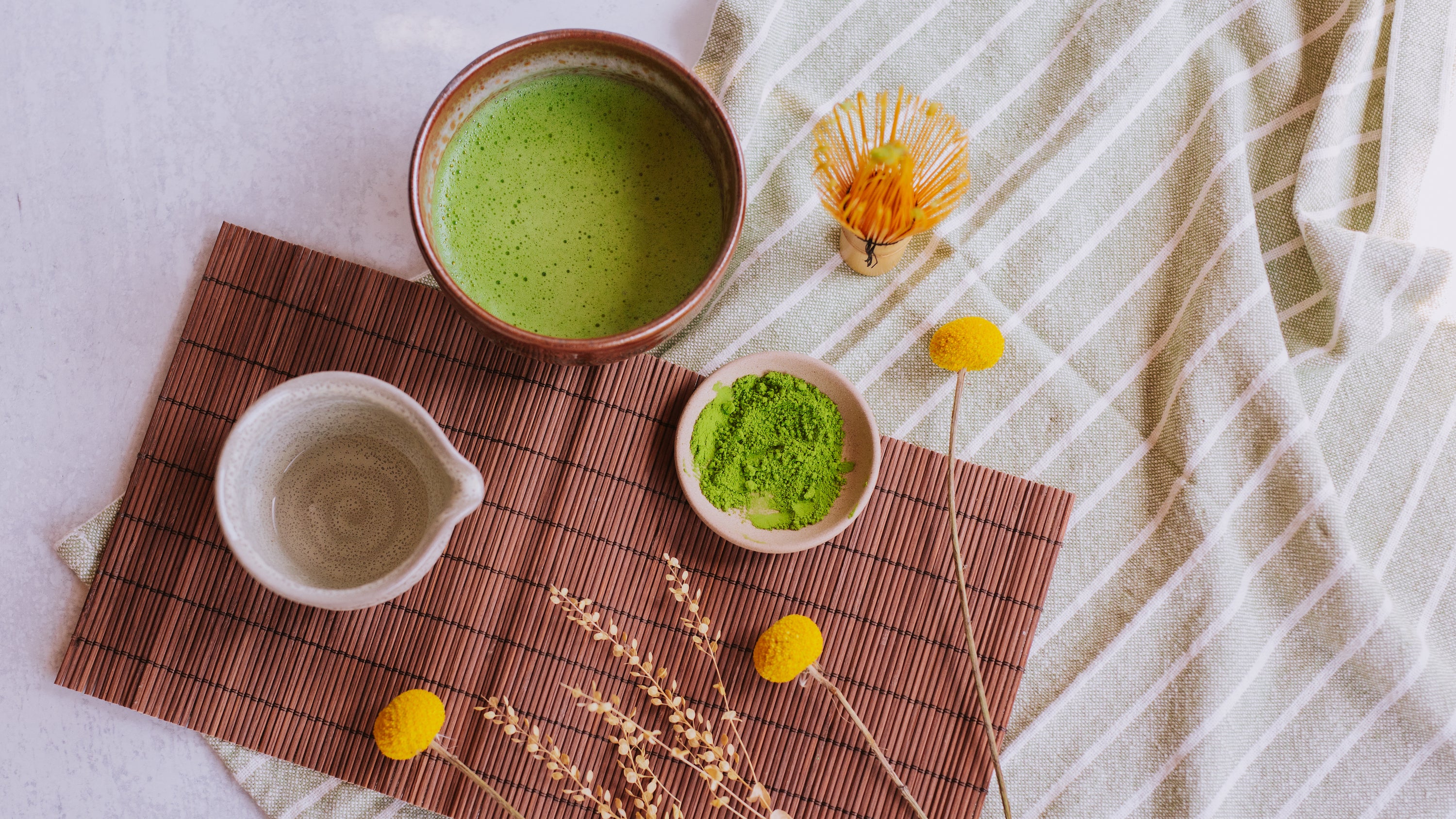 bowl of whisked matcha next to a matcha whisk, dish of matcha powder, and small pitcher of water