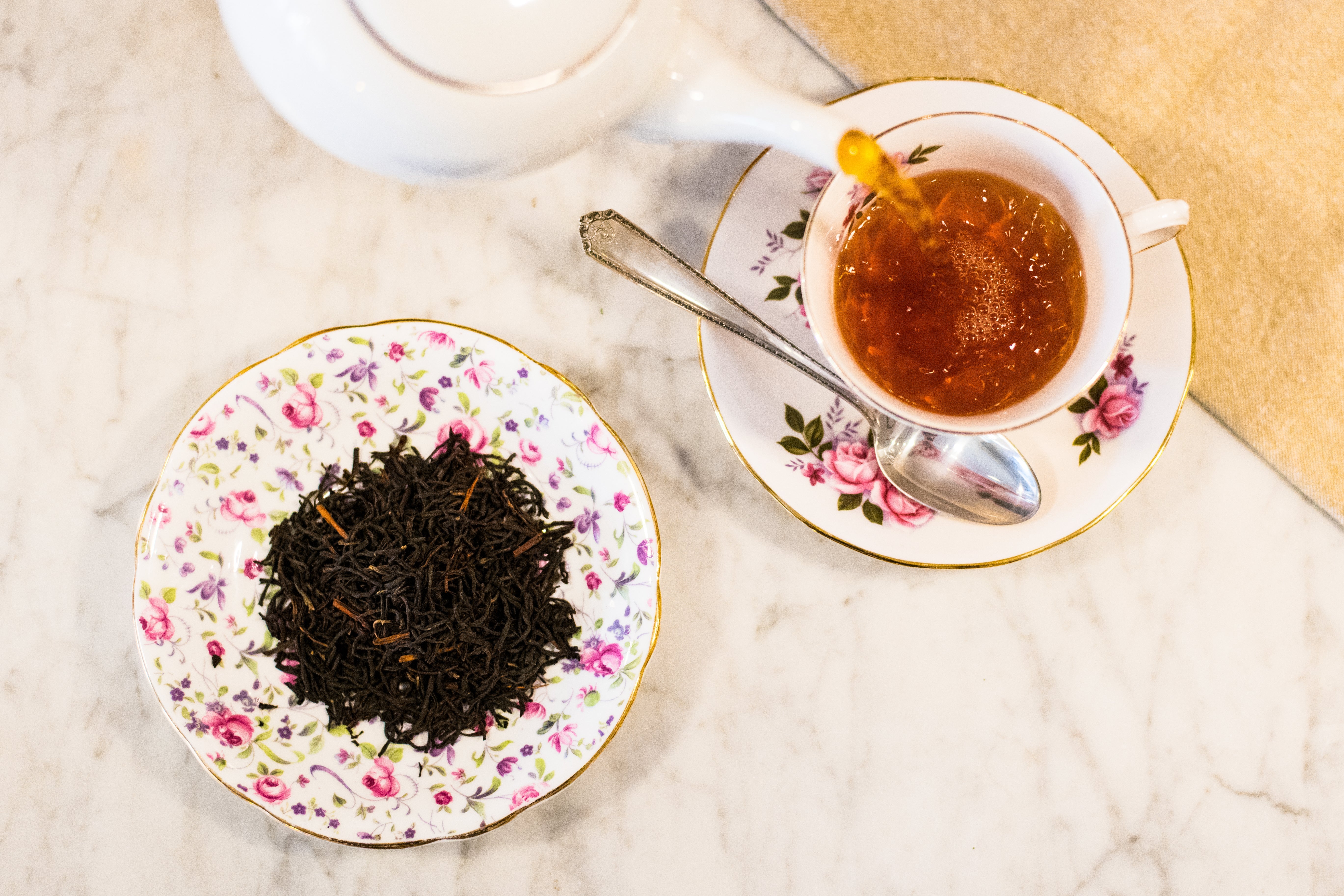 white teapot pouring black tea into a white and pink floral vintage teacup next to a small vintage dish of dry, loose leaf black tea