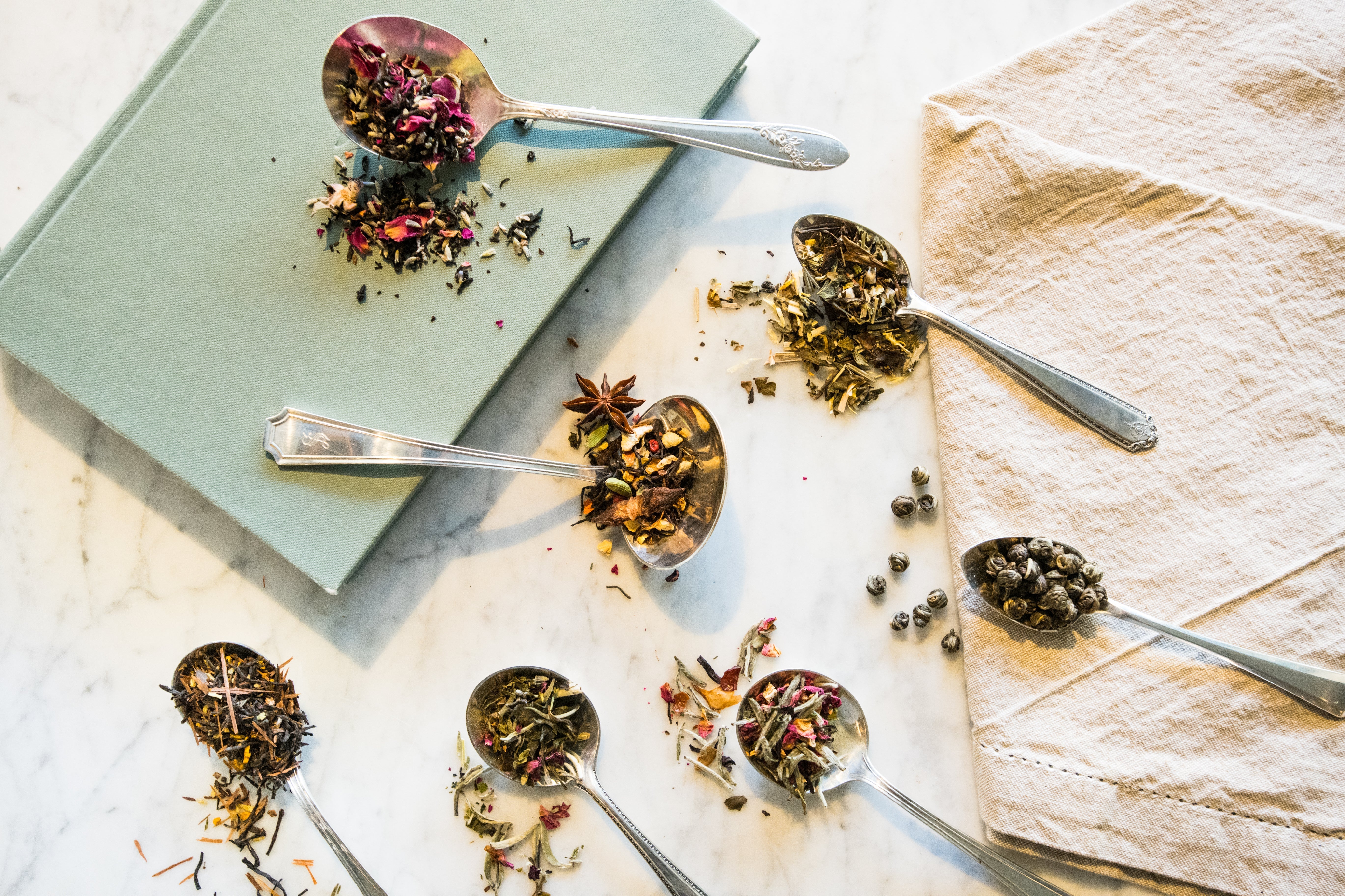 spoons of tea blends laid out in a random pattern on top of a book and linen napkin