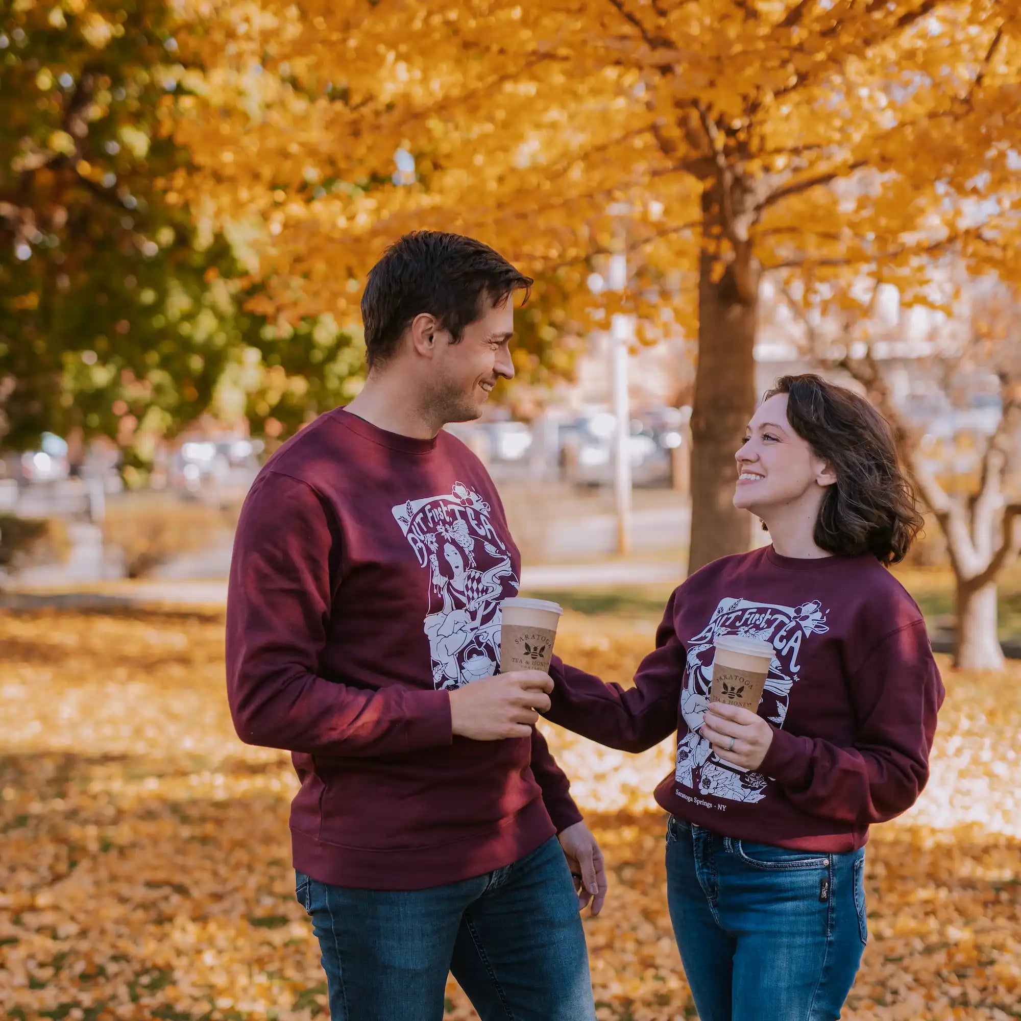 male-female couple wearing plum "but first, tea" sweatshirts and holding saratoga tea and honey to go tea cups in the fall