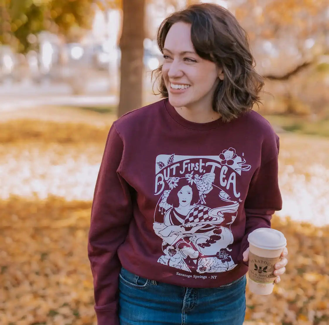 woman wearing maroon sweatshirt reading "but first tea" and holding a Saratoga Tea & Honey Co. to-go cup