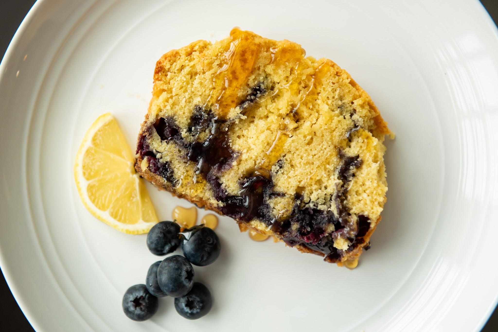 slice of lemon ginger blueberry bread on white plate with fresh berries and lemon, all drizzled with honey