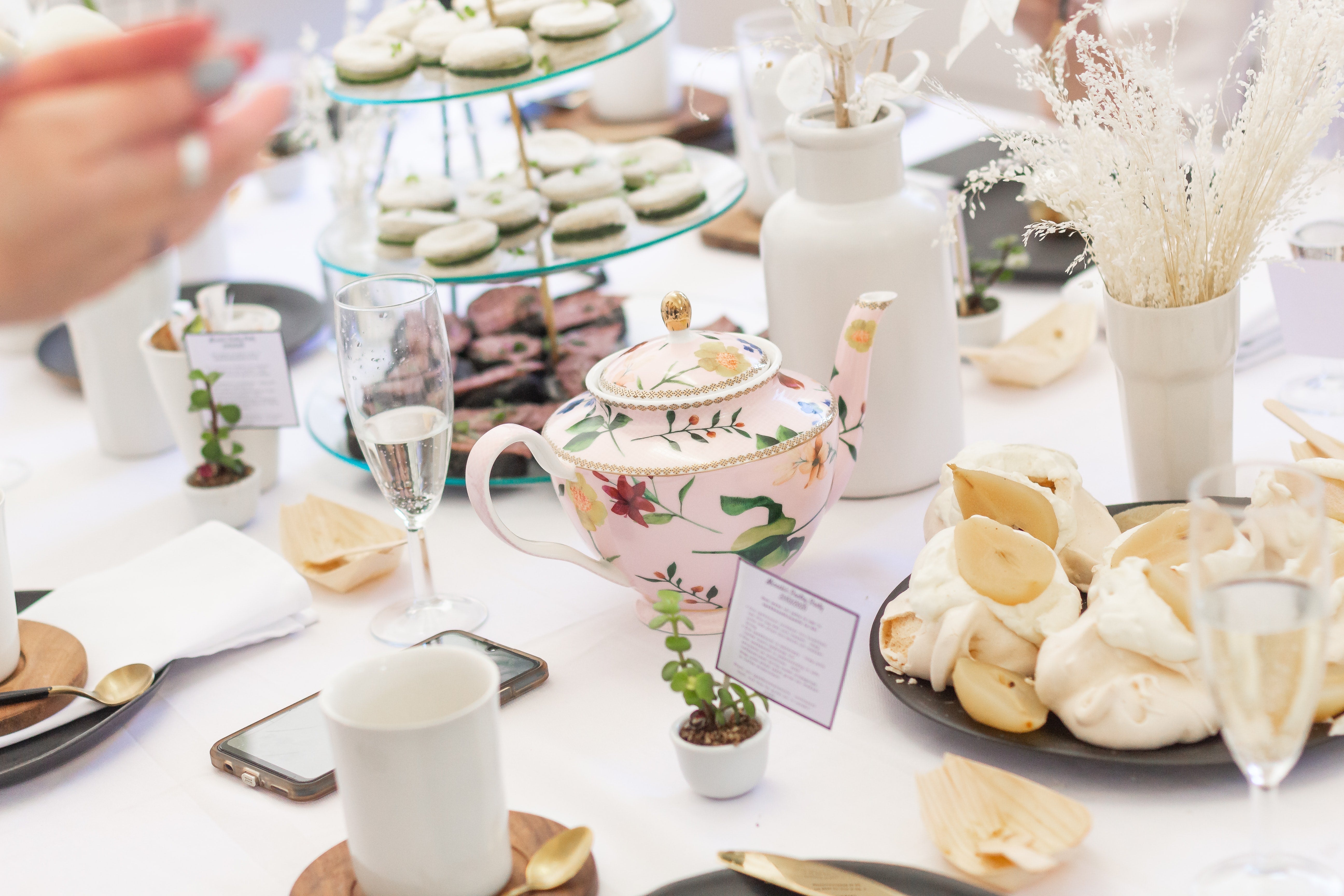 table set for afternoon tea with a vintage floral teapot, three tier stand with macarons, flowers, and other sweet treats