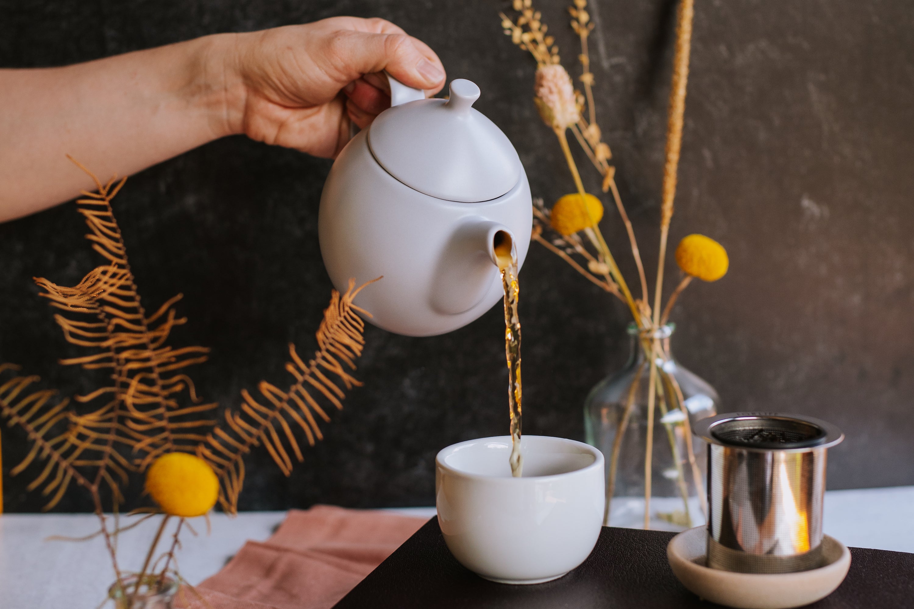 hand pouring hot tea from teapot into cup with tea infuser sitting to the side